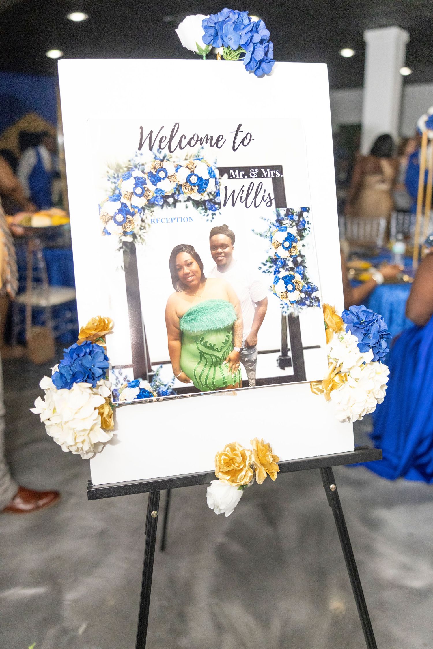 Wedding welcome sign for Mr. & Mrs. Willis on easel, decorated with blue, white, and gold flowers with couple photo.