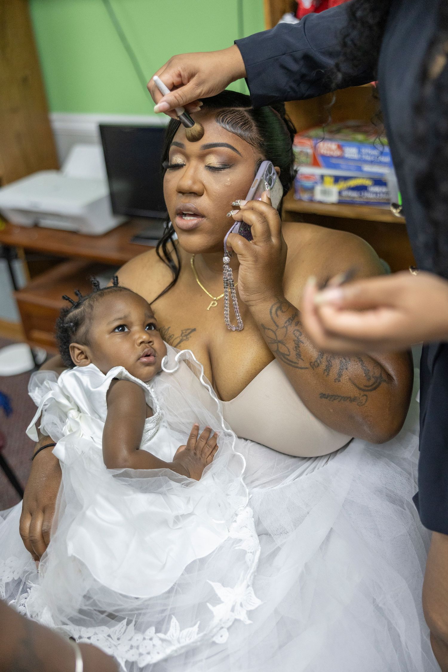 A bride holds her baby girl while getting hair done and talking on phone before her wedding ceremony.