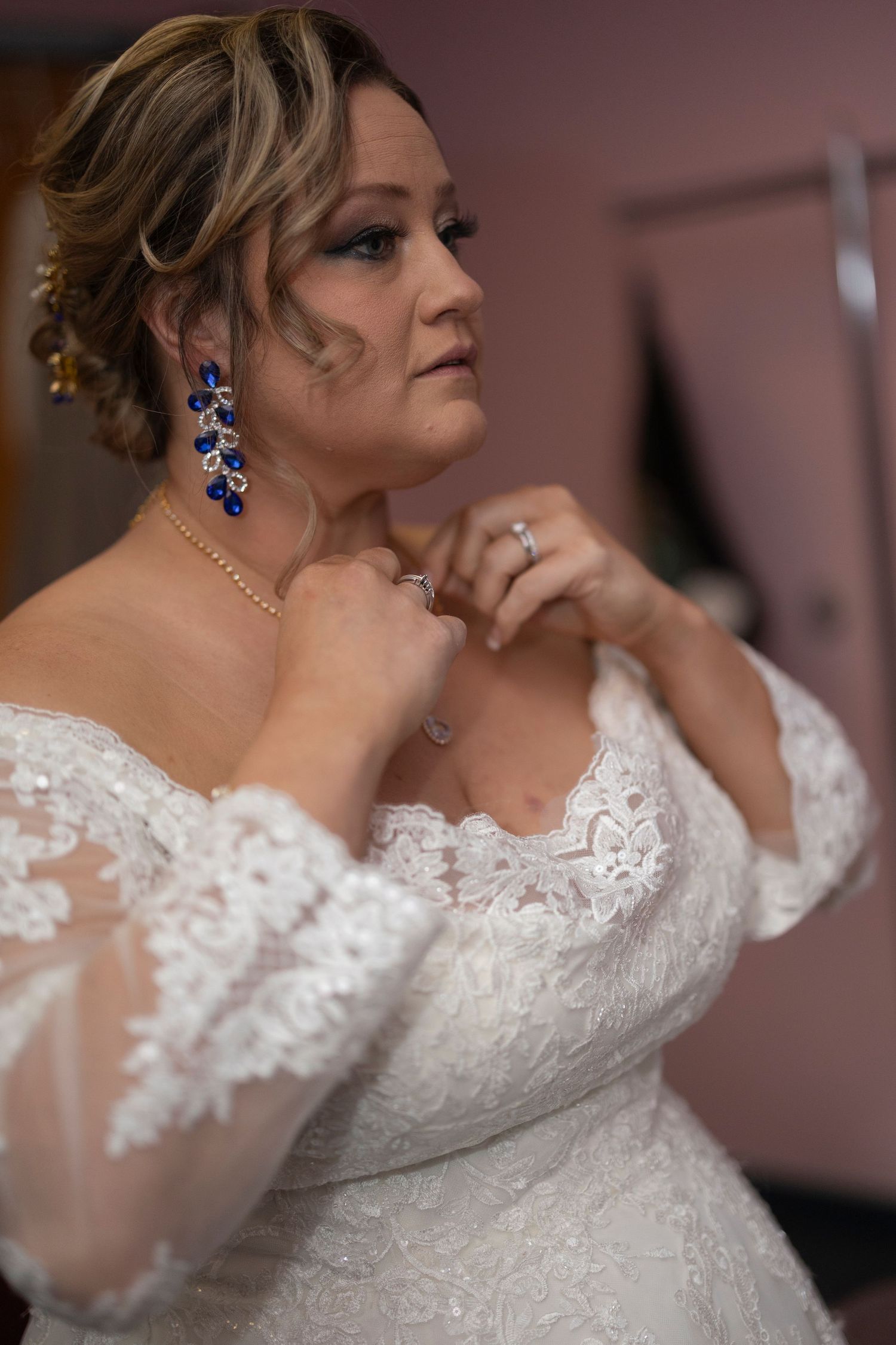 Bride in off-shoulder lace wedding gown adjusting necklace, wearing blue drop earrings and diamond ring.