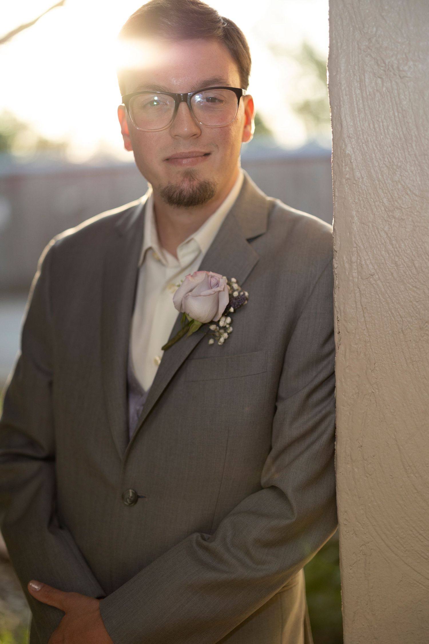 A groom in a grey suit and glasses poses outdoors wearing a pink rose boutonniere.