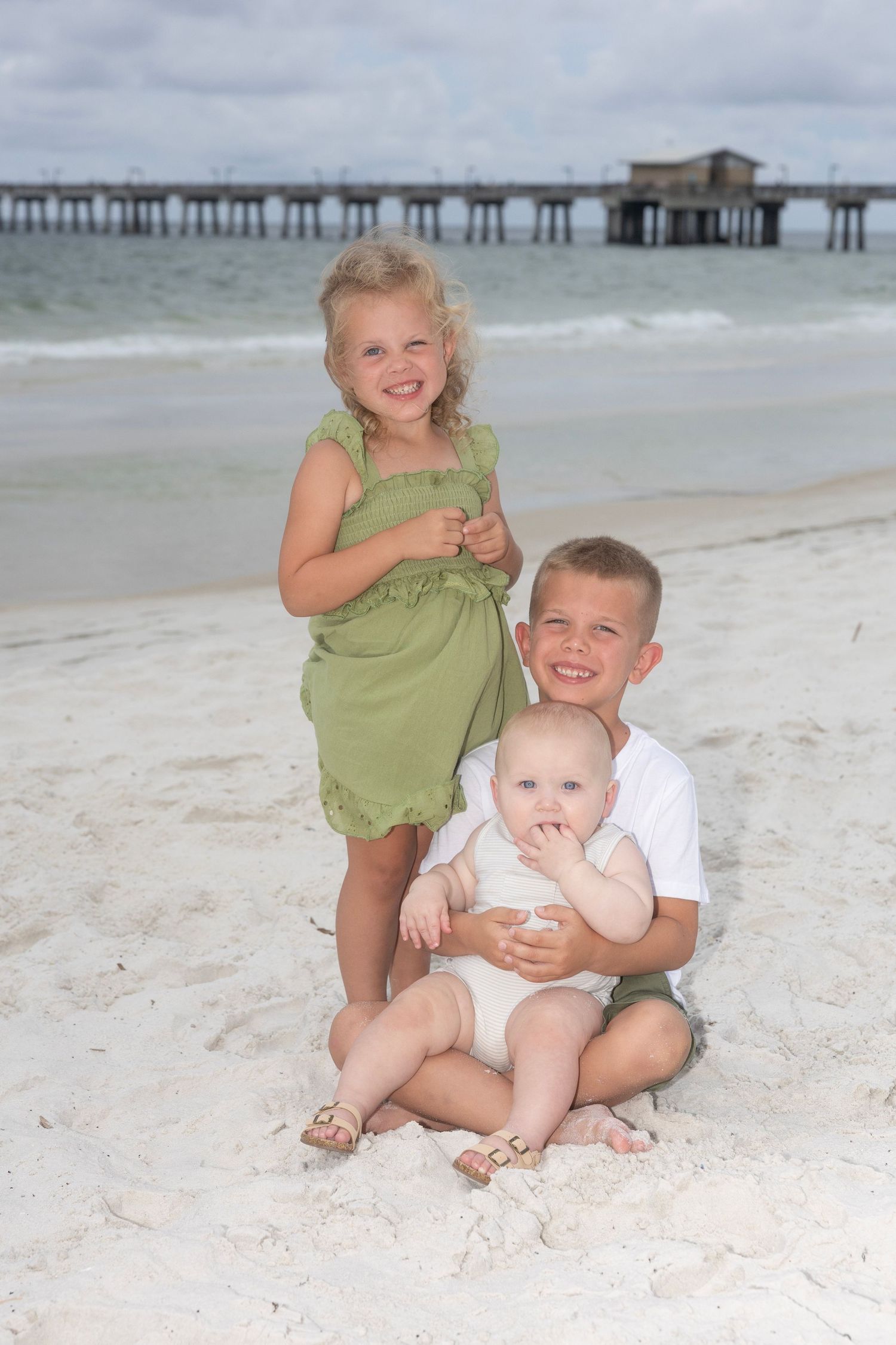 Three children sitting together on a beach with a wooden pier visible in the background.