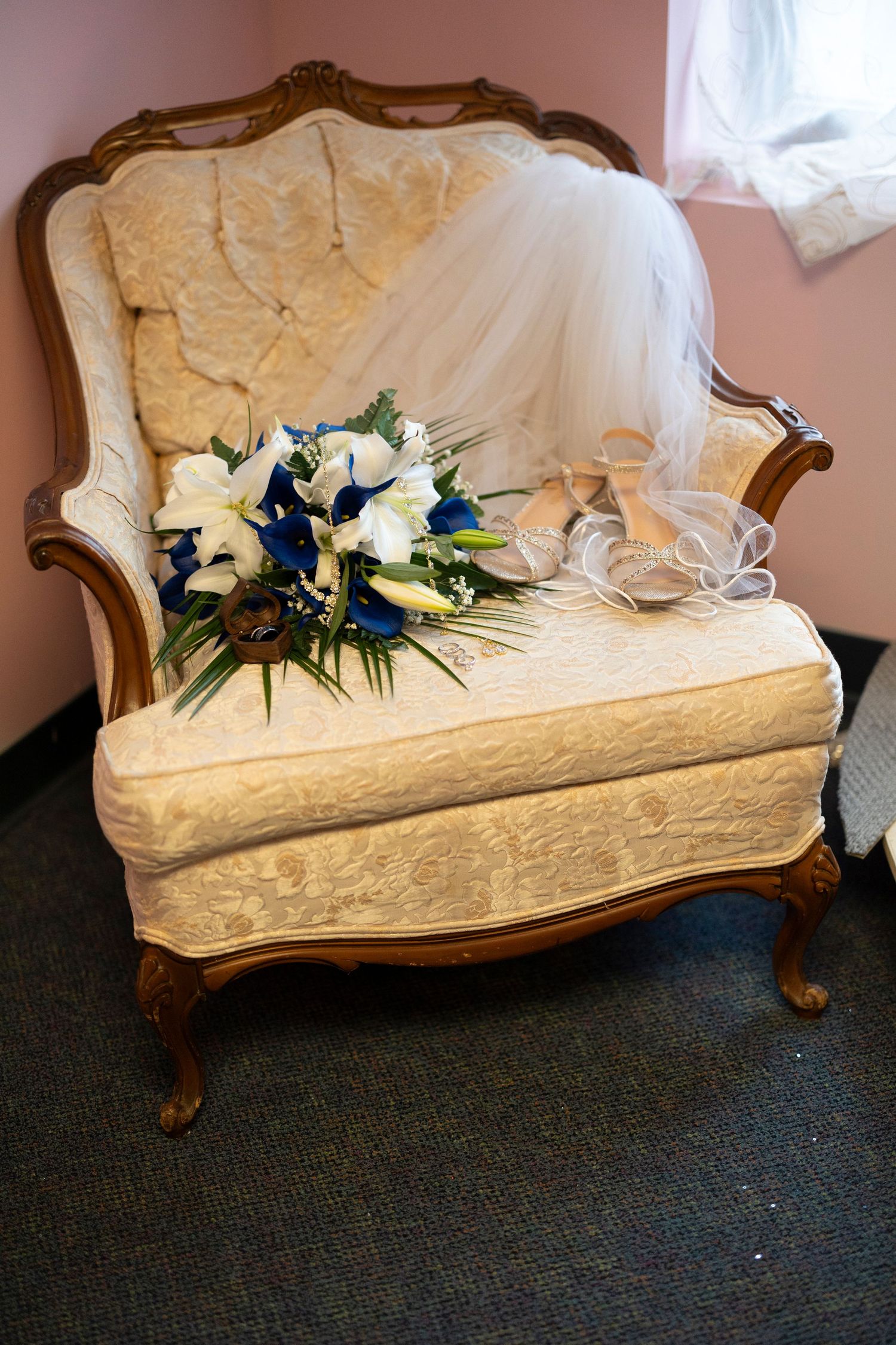 Elegant vintage cream armchair displaying a bridal bouquet of blue and white flowers, wedding veil, and heeled shoes.