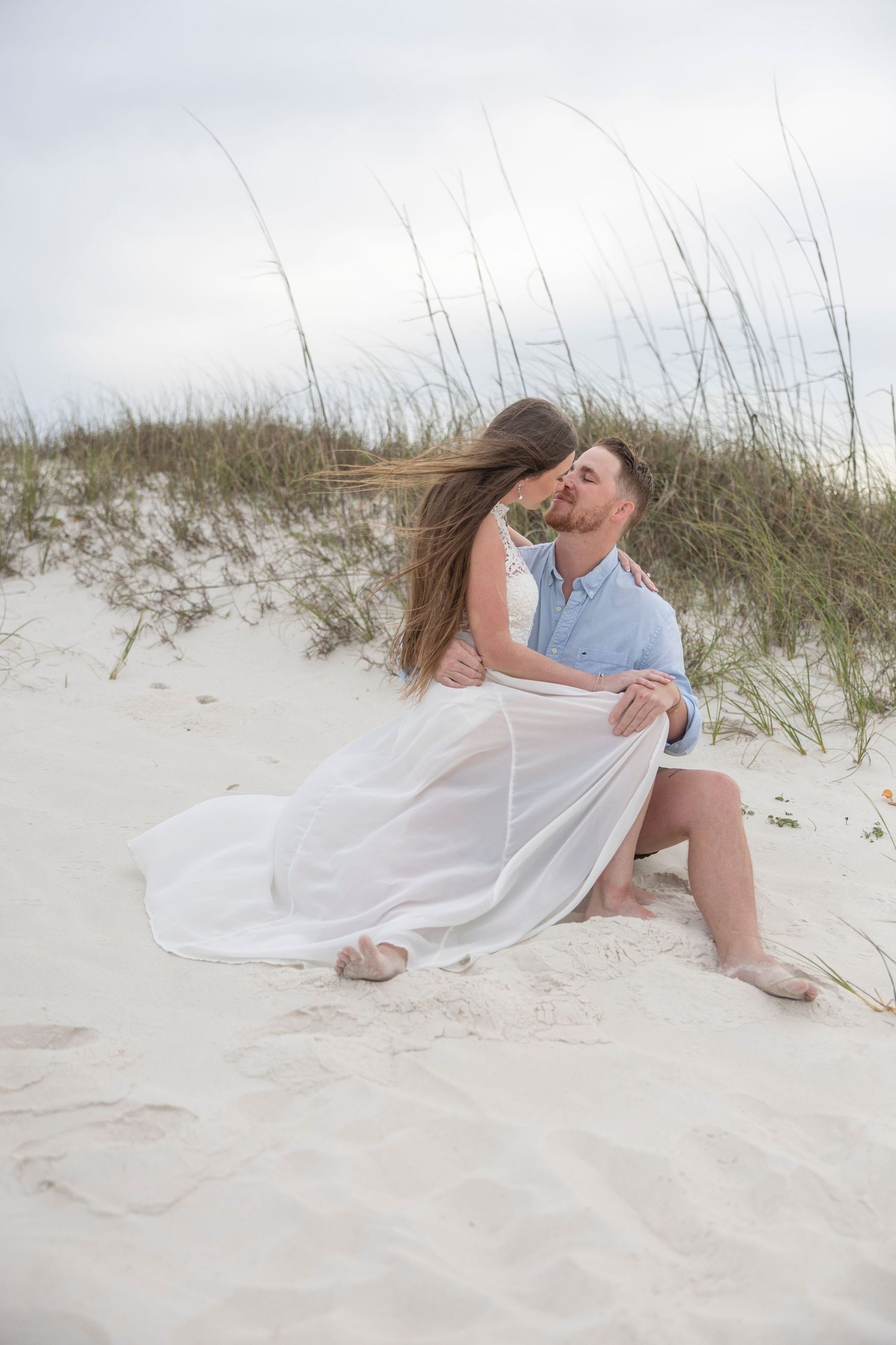 Couple kissing on sandy beach dunes, woman in flowing white dress, man in blue shirt, romantic outdoor engagement photo.