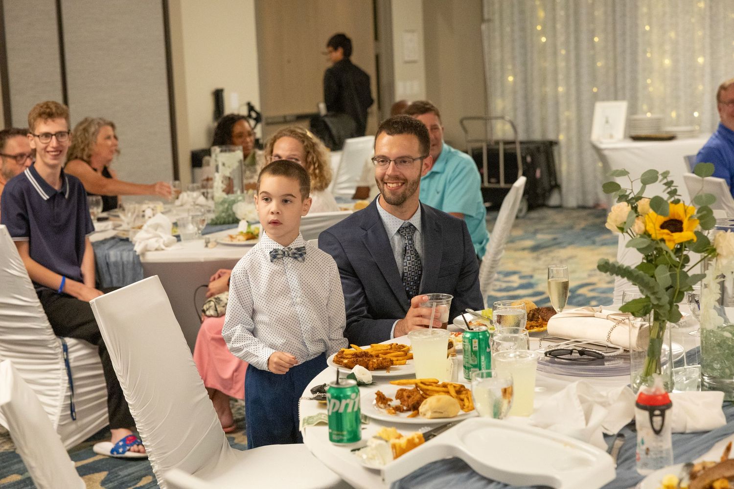 A man and young boy in bow tie smile at a wedding reception dinner table with sunflowers and food.