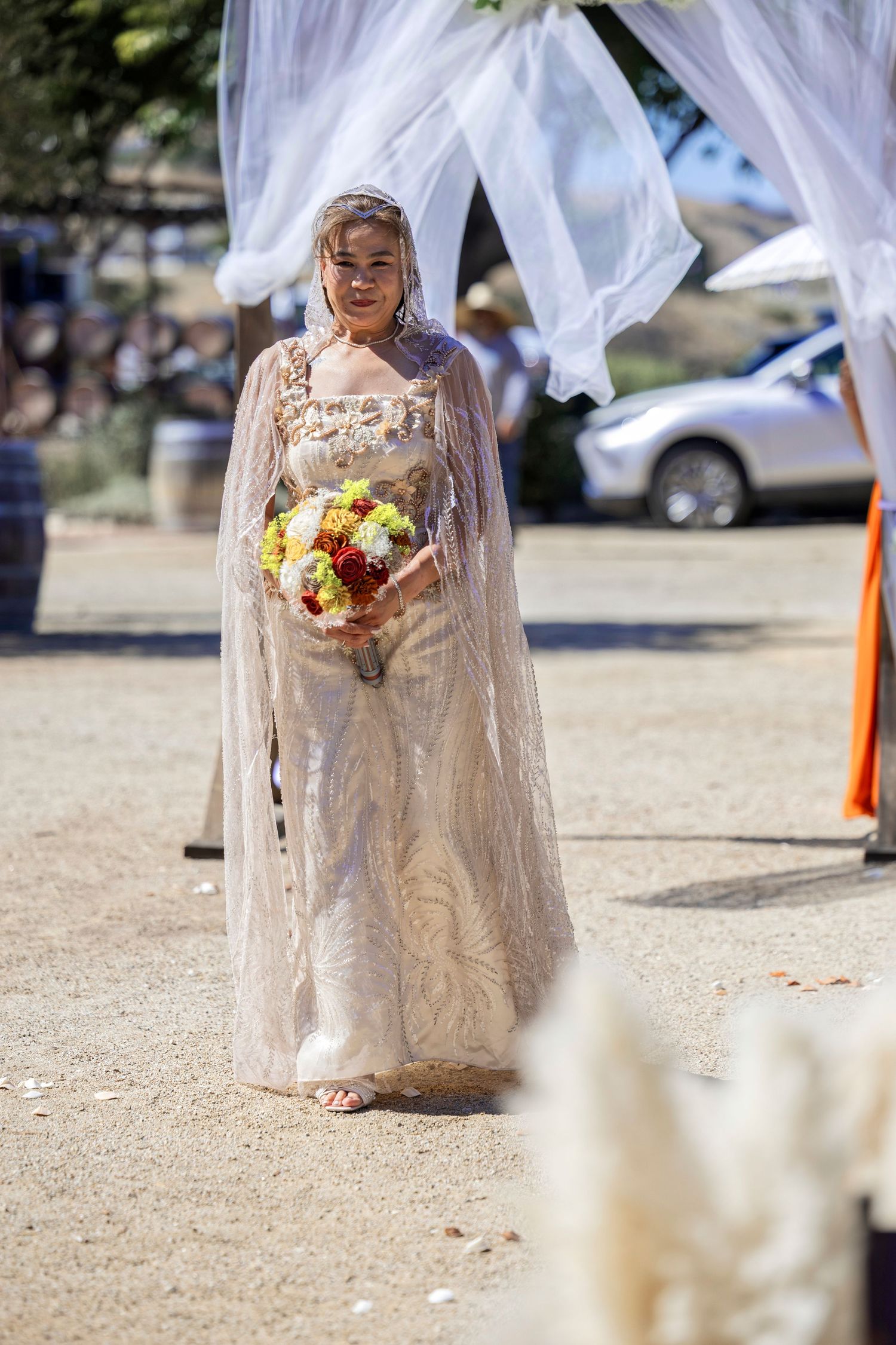 Smiling bride in lace gown and veil holds colorful bouquet outdoors at sunny wedding ceremony.