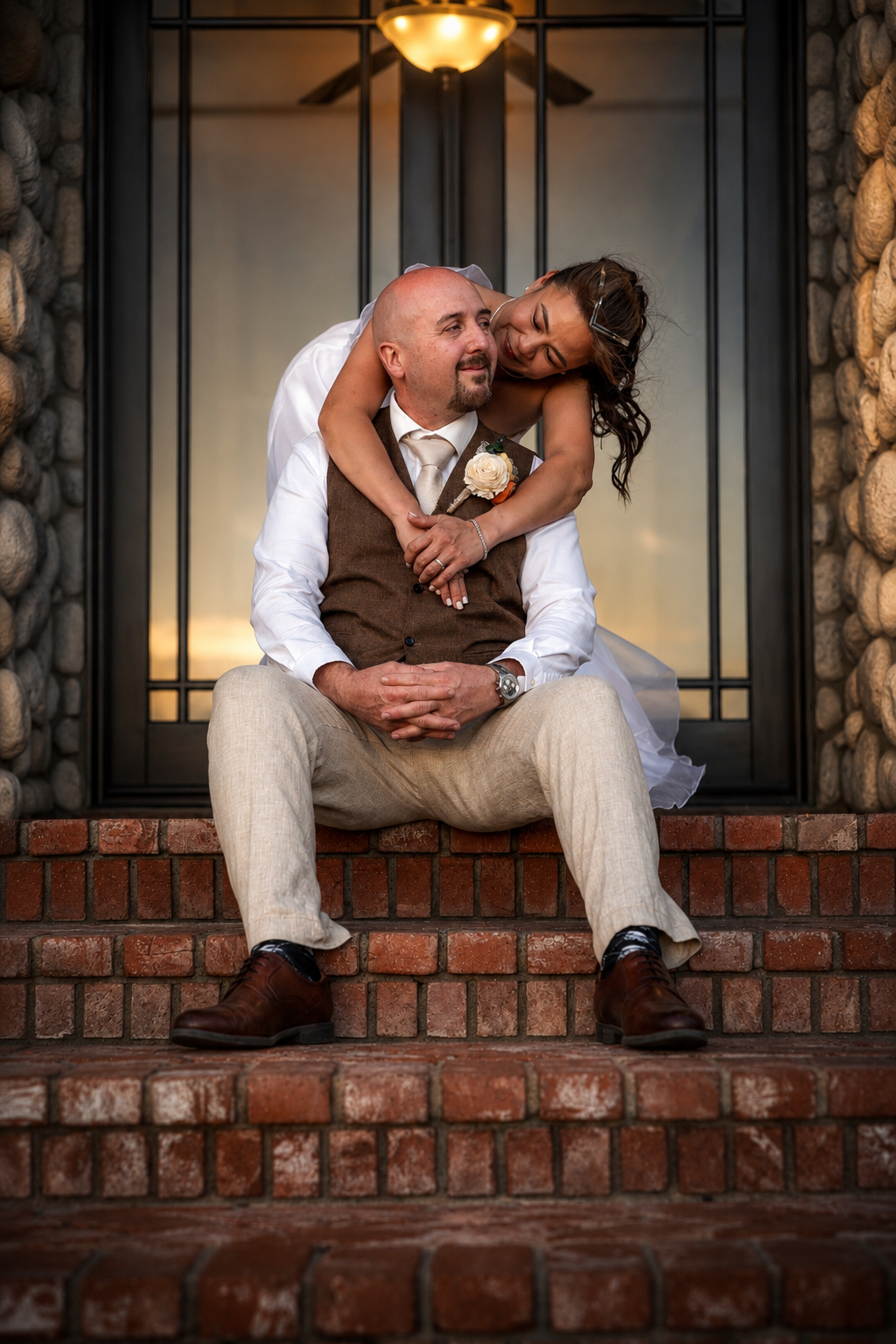 A bride embraces her groom from behind as they sit on brick steps outside a stone building during their wedding.