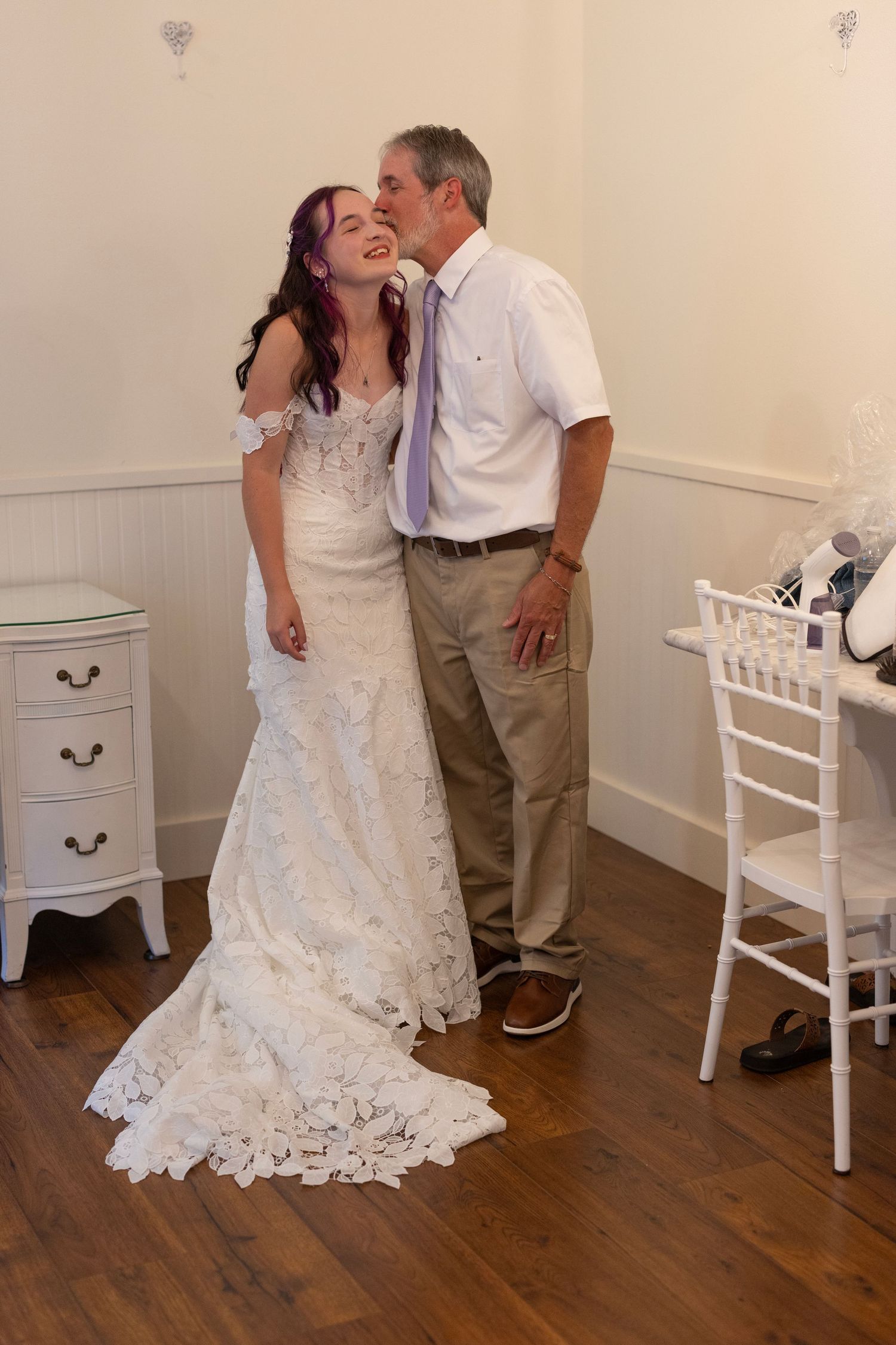 Bride in lace wedding gown shares a tender moment with older man in white shirt and khakis at indoor wedding venue.