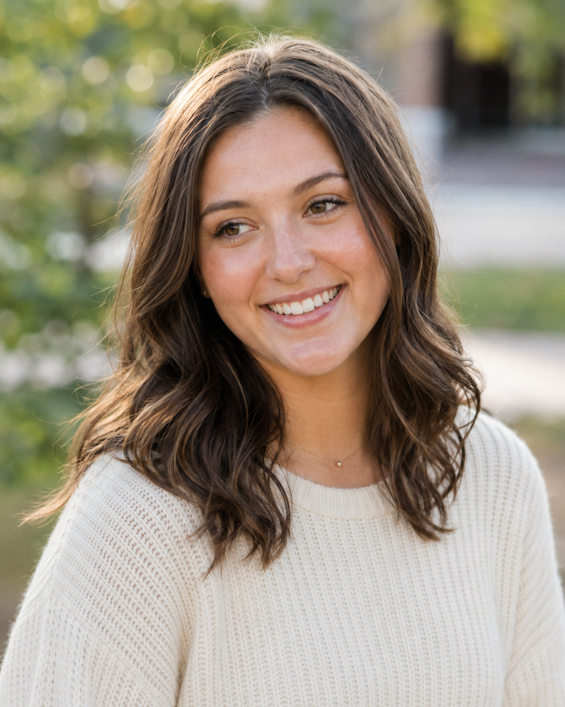 Smiling young woman with wavy brown hair wearing a cream sweater outdoors.