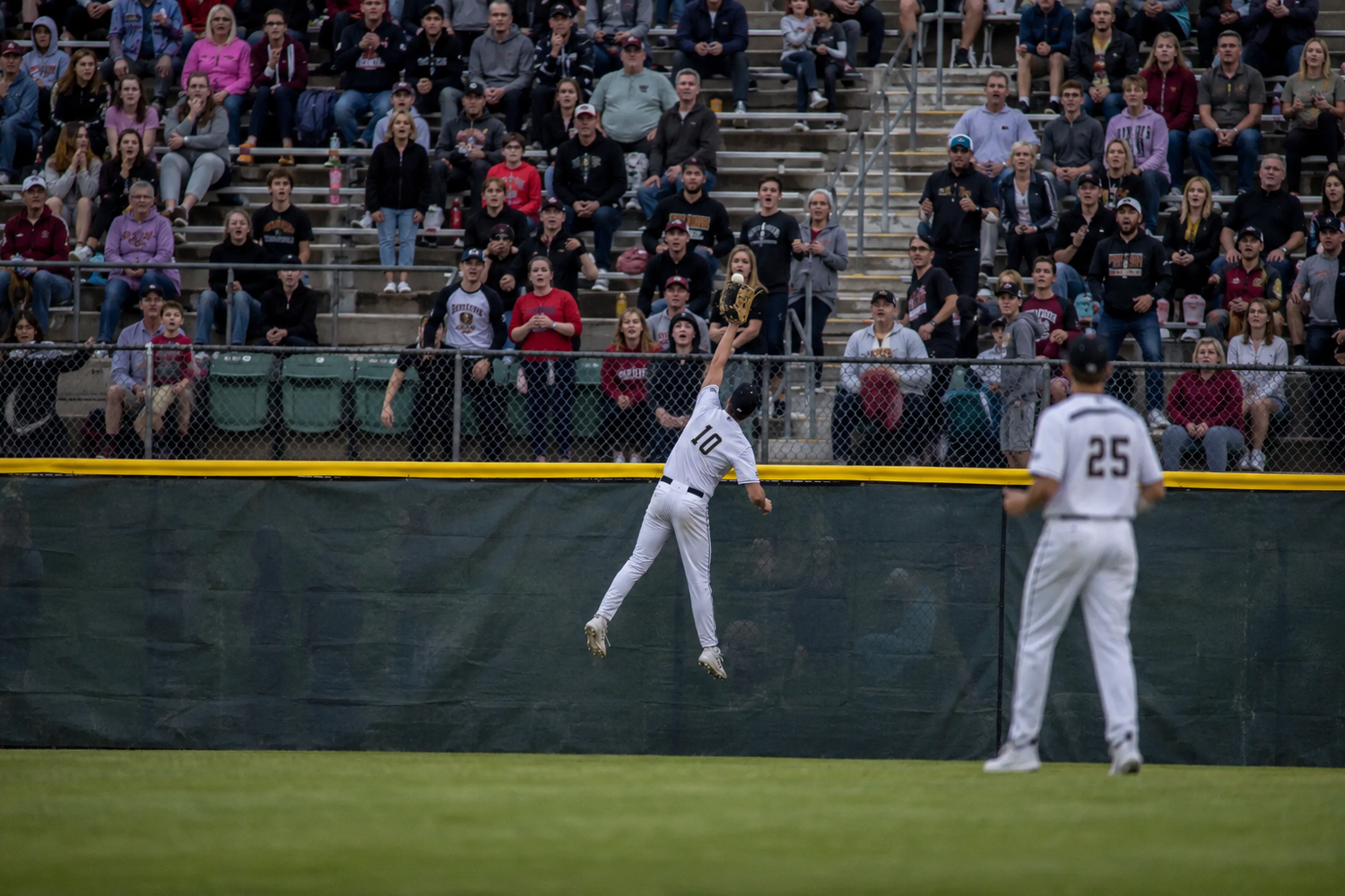 Outfielder leaps at the warning track wall attempting to catch a fly ball before a packed crowd.