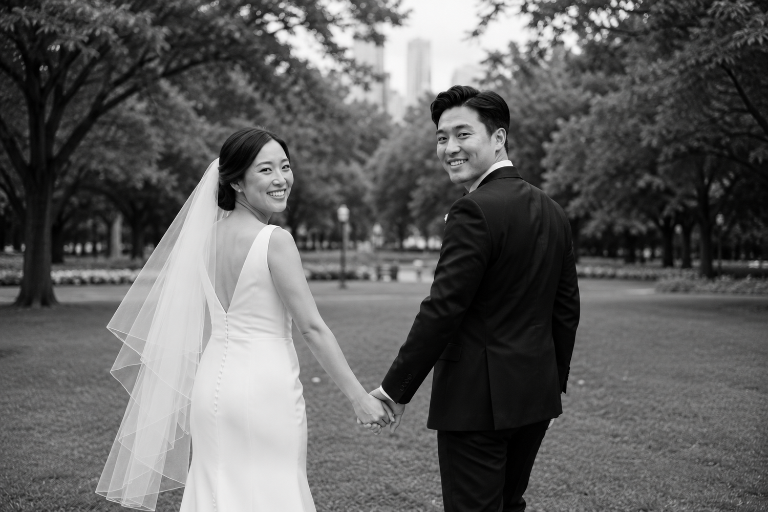 Black and white photo of smiling Asian bride and groom holding hands walking through a park on their wedding day.