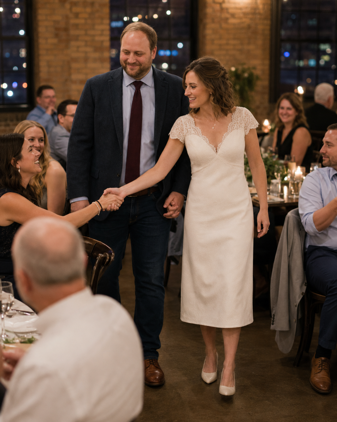 Bride in white dress and groom in navy suit greet wedding guests at elegant reception venue.