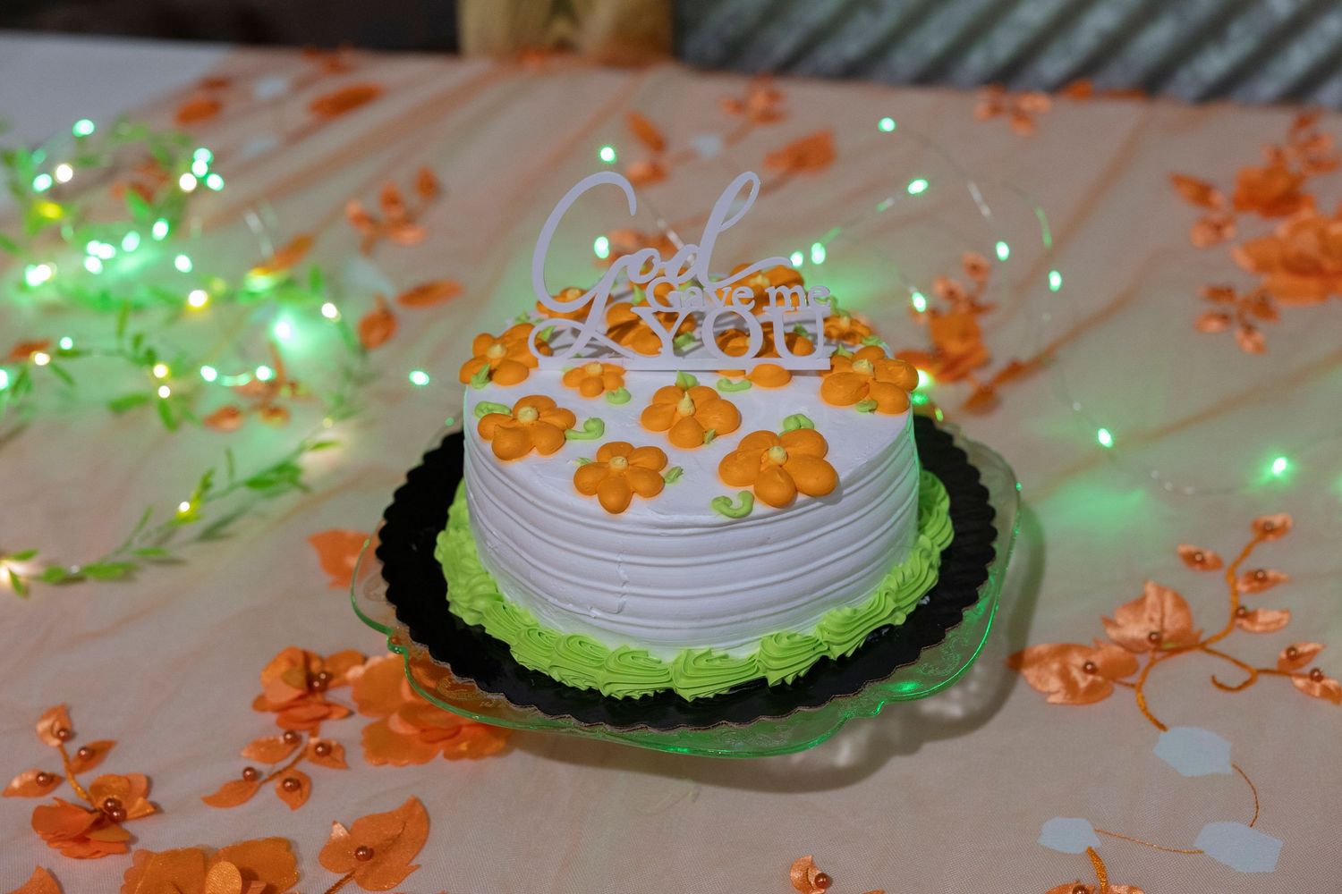 White cake with orange flowers and fairy lights on a decorated table.