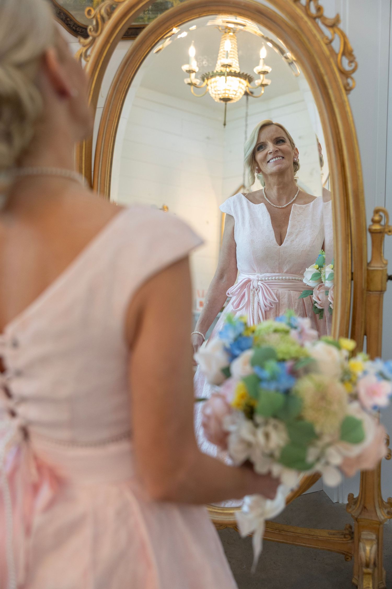 Bride in white dress holding colorful bouquet smiles at her reflection in ornate gold mirror, bridesmaid in pink nearby.
