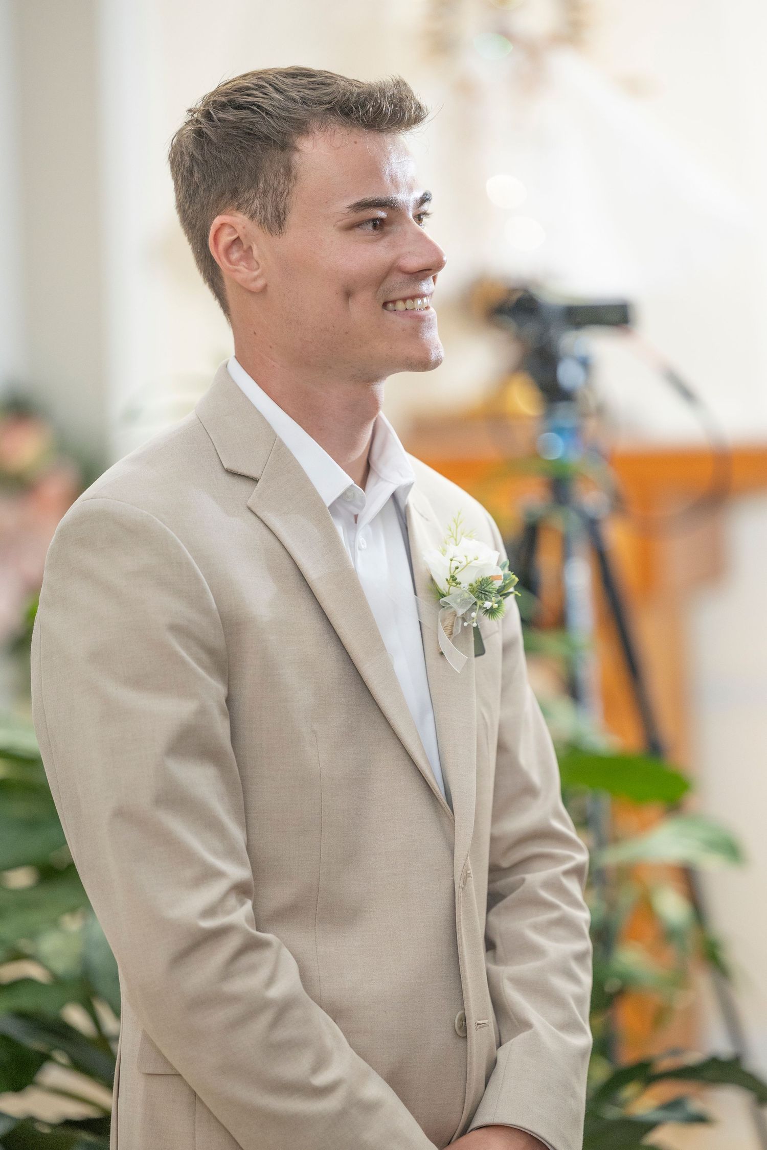 A groom in a tan suit and white shirt smiles during a wedding ceremony.