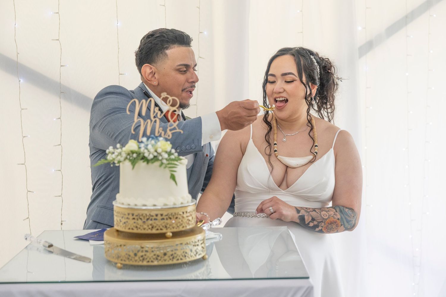A couple laughs together while cutting their gold-colored wedding cake in a bright white room.