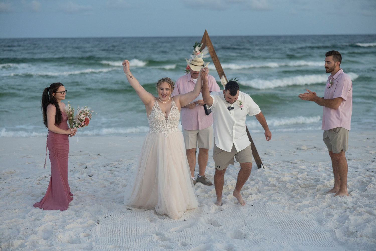 A couple celebrates their beach wedding ceremony with loved ones on pristine white sand at sunset.