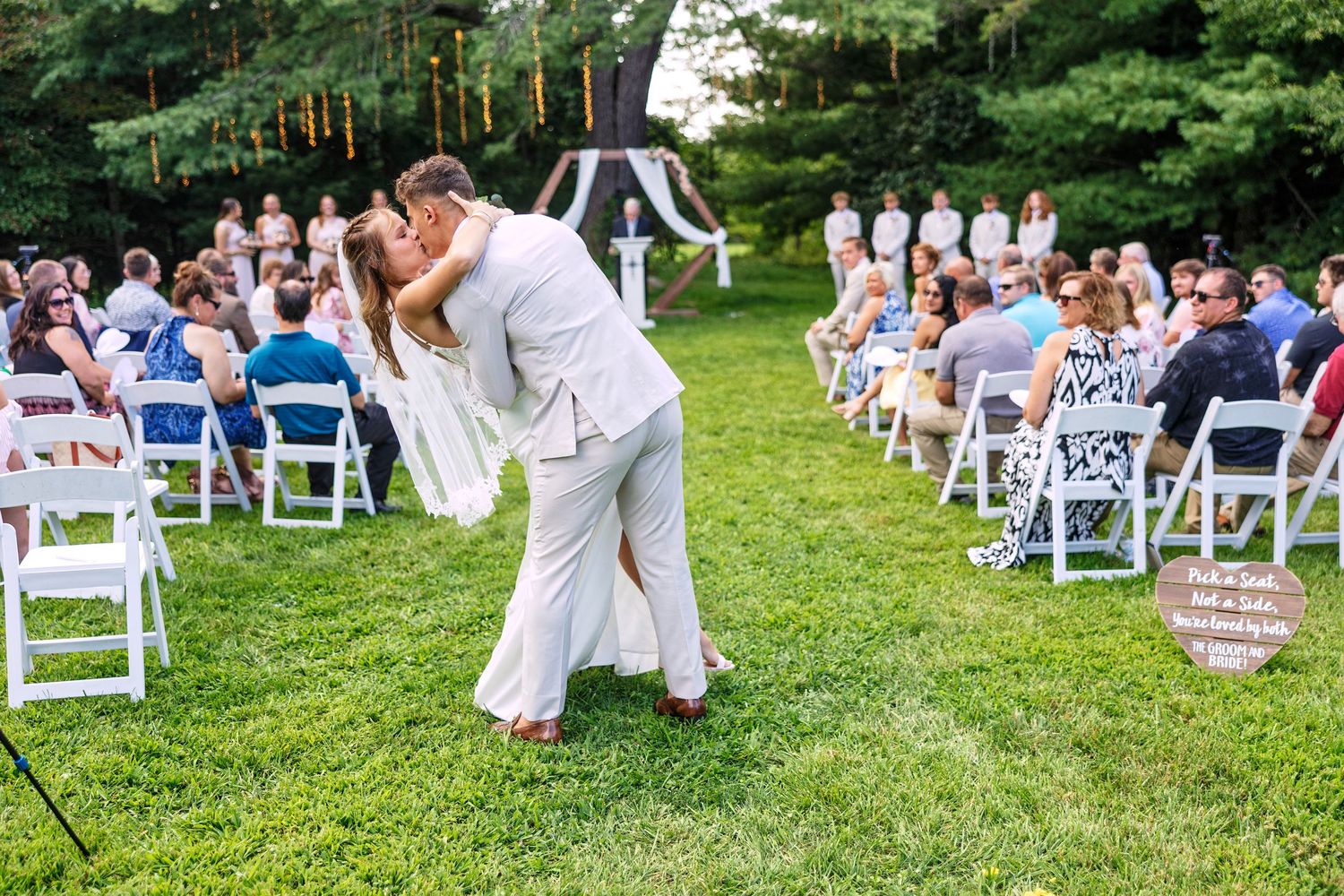 Panoramic view of an outdoor wedding ceremony setup with white chairs on a lush green lawn surrounded by trees.
