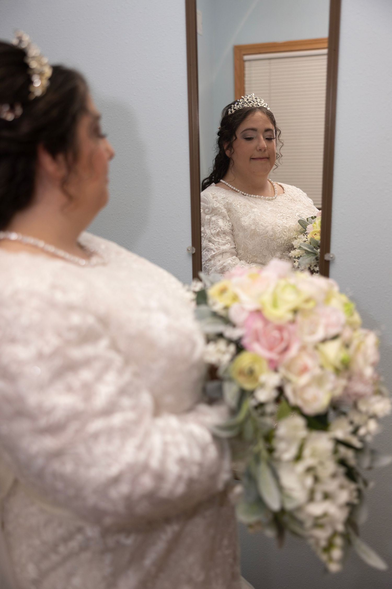 A bride in a white lace dress holds a pastel bouquet of roses and flowers while getting ready for her wedding ceremony.