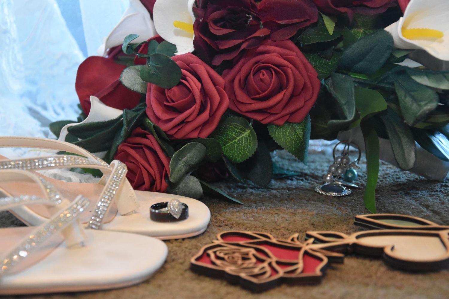 Deep red roses with green foliage arranged alongside elegant white bridal shoes and delicate accessories.