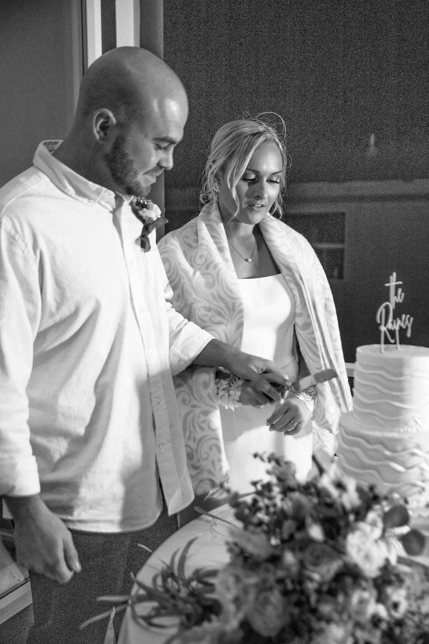 A couple cuts their elegant white wedding cake together while standing next to floral decorations in black and white.