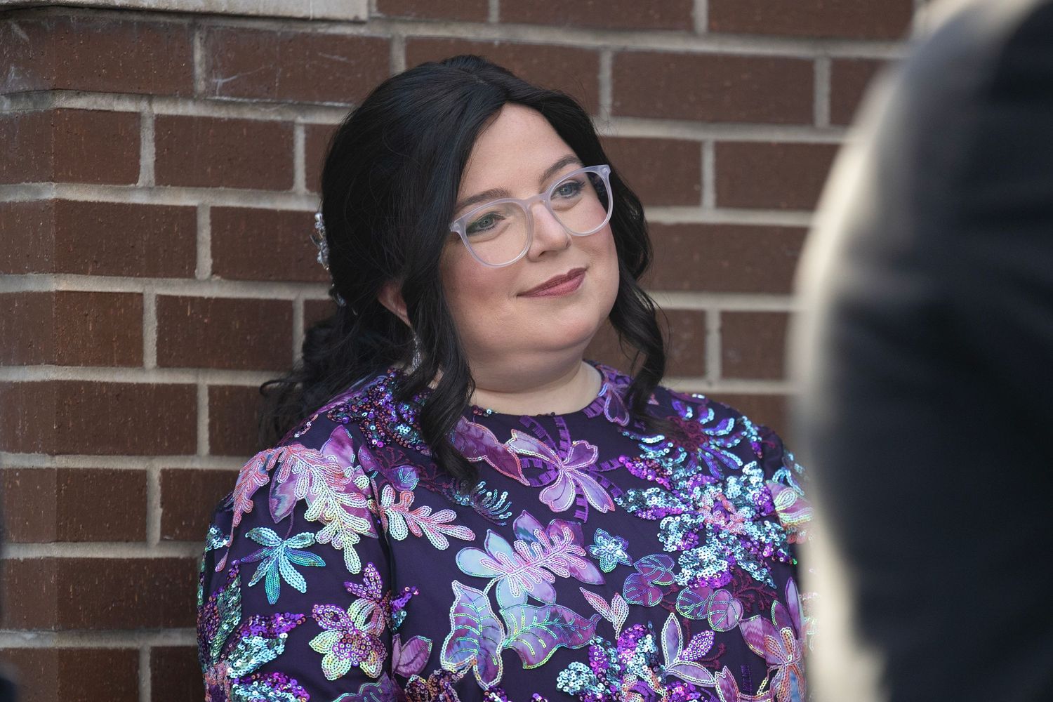 Someone wearing glasses and a floral print dress with purple and blue flowers stands against a brick wall.