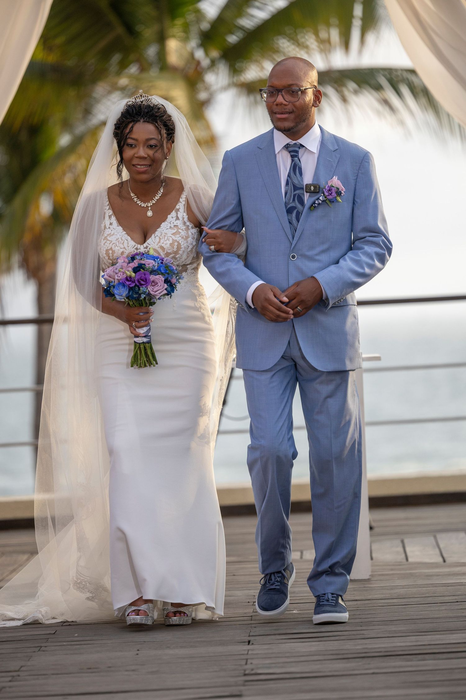 A wedding couple stands on a wooden deck with palm trees in the background during a tropical beach ceremony.