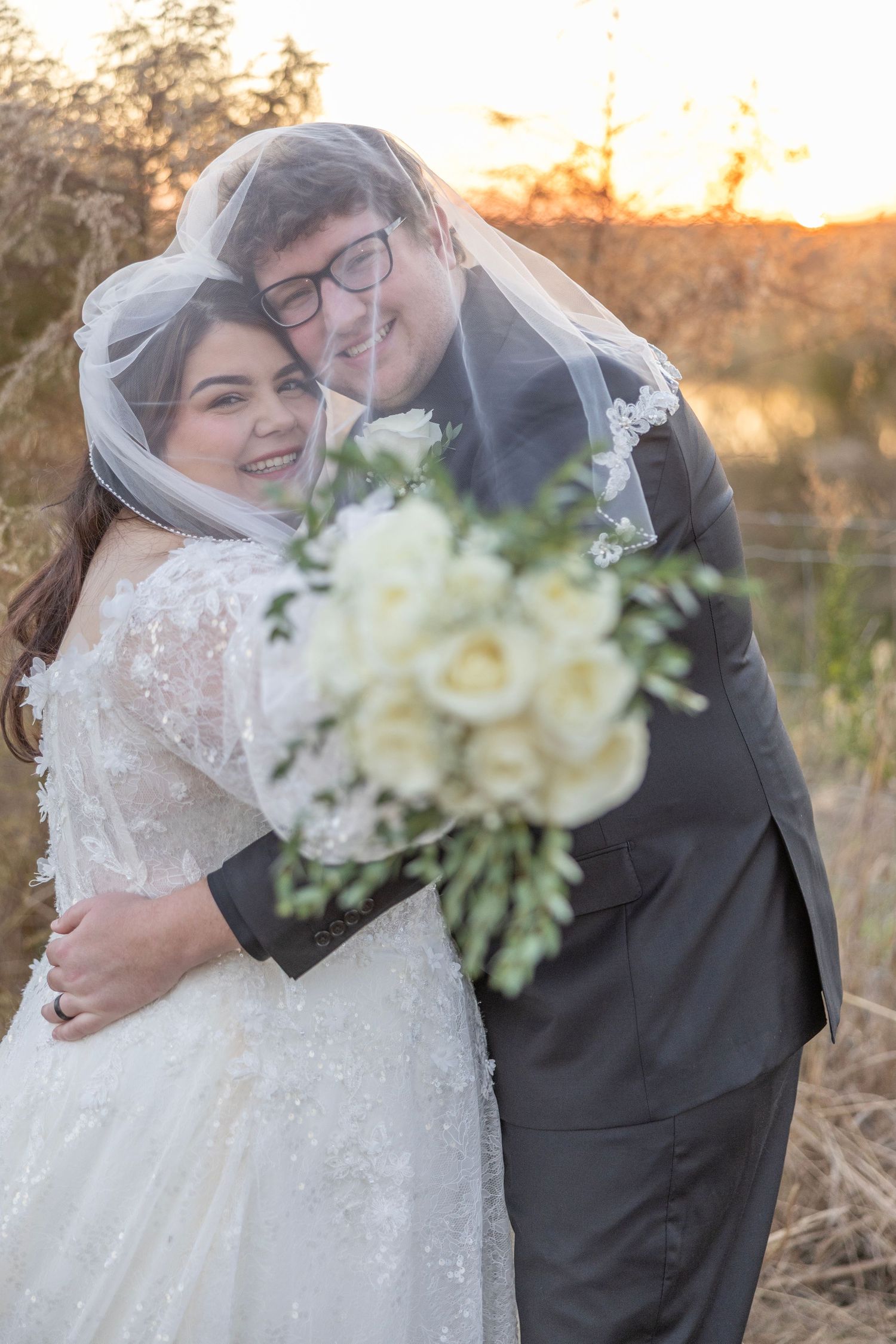 Romantic newlywed couple embraces during golden hour sunset in a rustic outdoor setting.