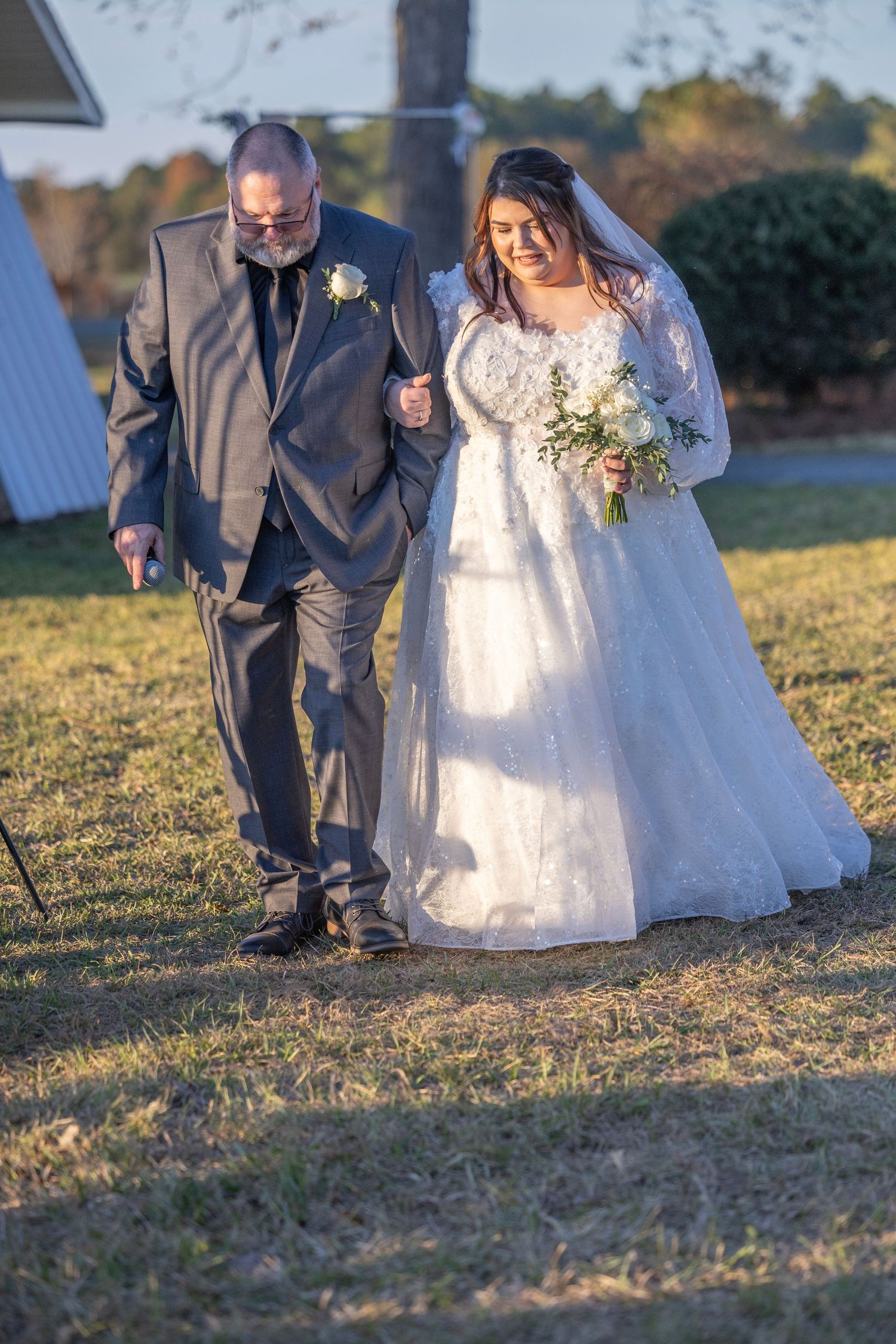 A bride in a flowing white lace gown and groom in gray suit walk together on sunlit grass during golden hour.