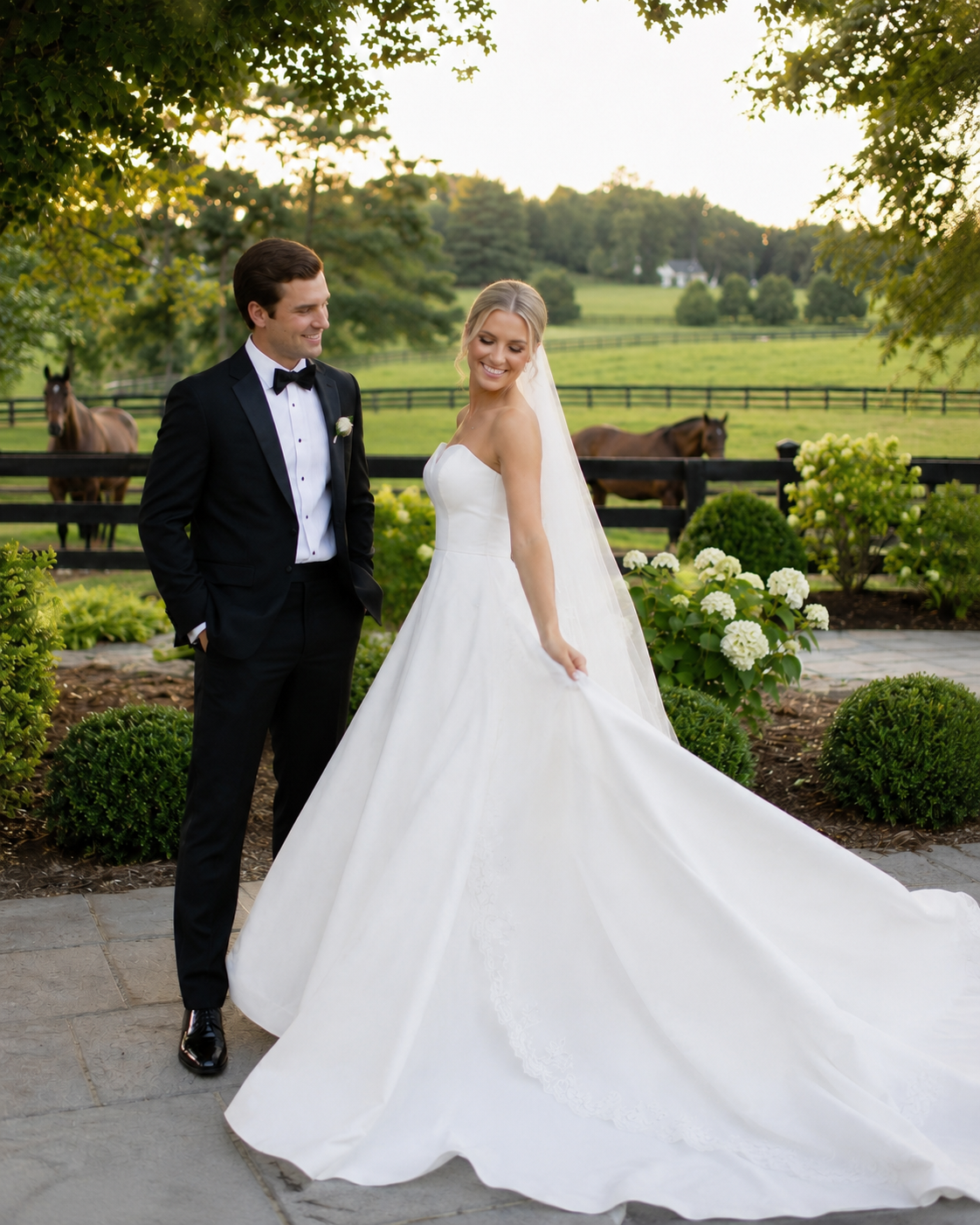 Bride in strapless ballgown and groom in tuxedo pose on garden path with horses and green fields backdrop.