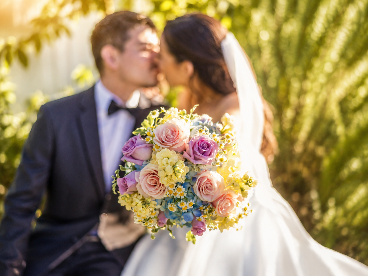 A bride and groom kissing while holding a colorful bouquet of pink roses and yellow flowers on their wedding day.