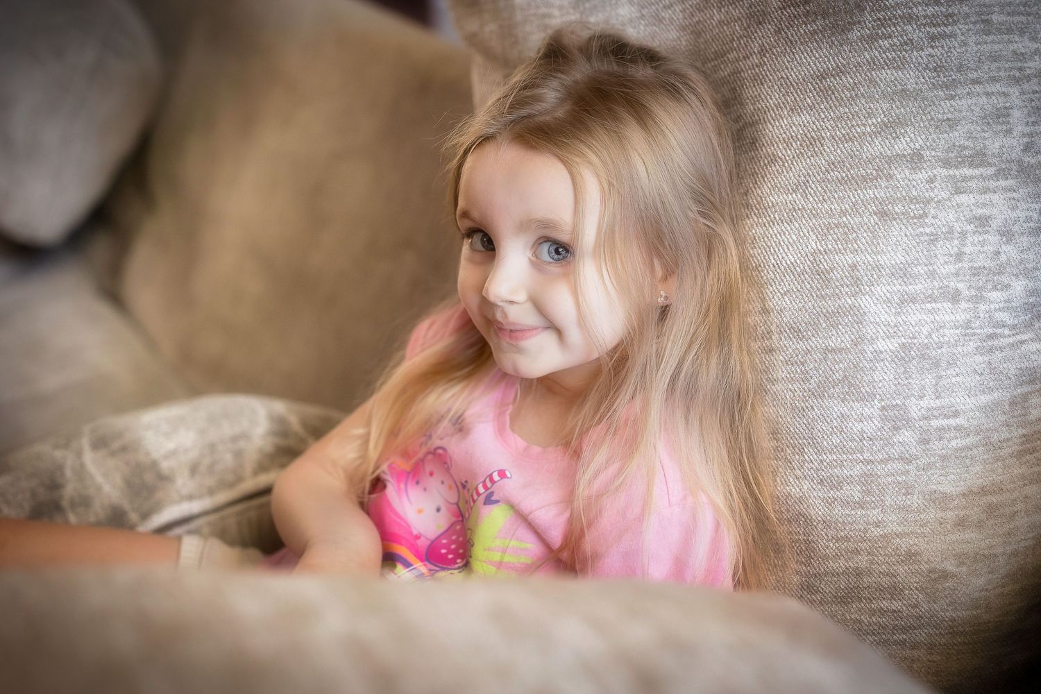 A young child in a pink dress sits on a gray couch while holding a stuffed toy.
