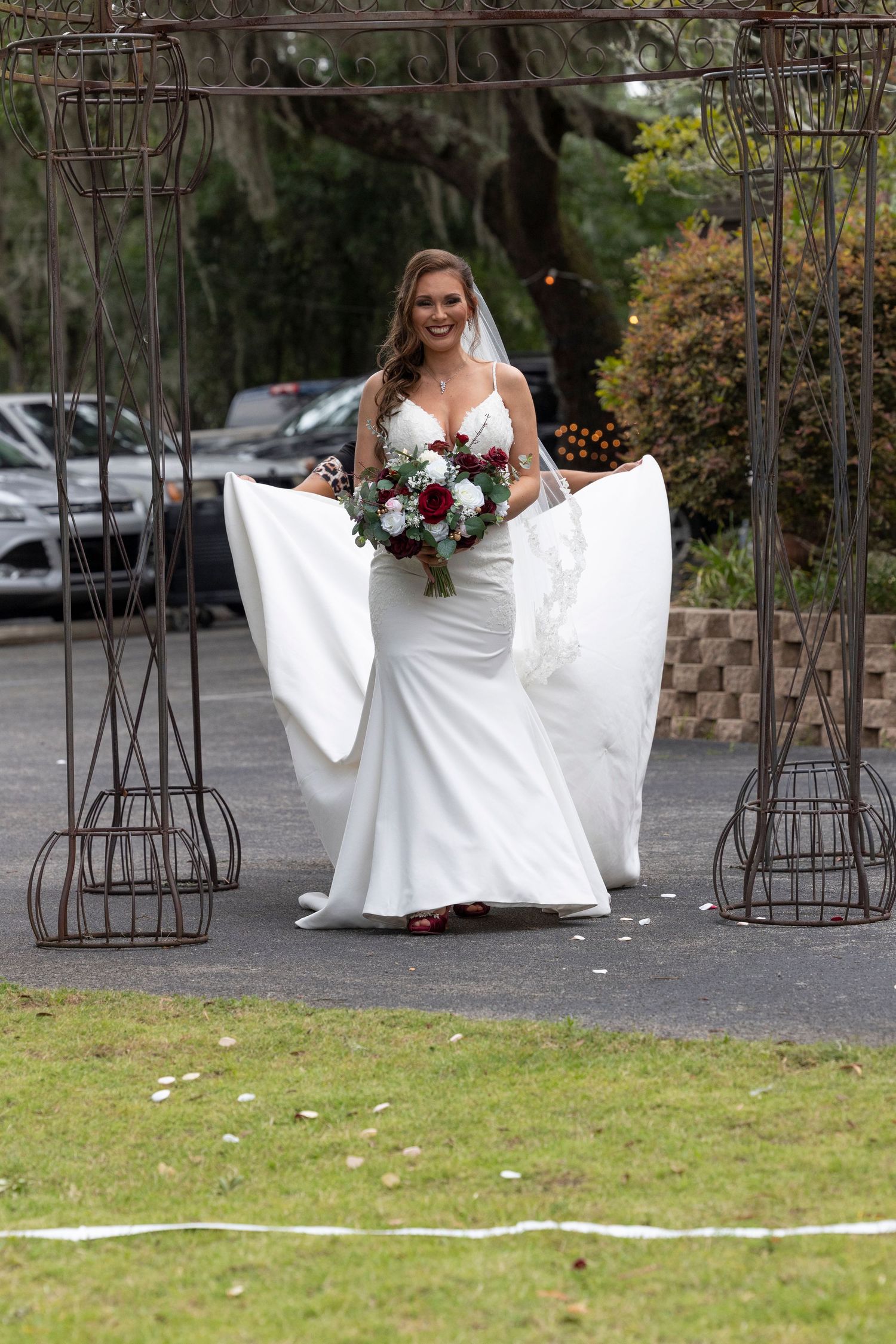 Beautiful bride in white gown standing between metal arches with burgundy bouquet.