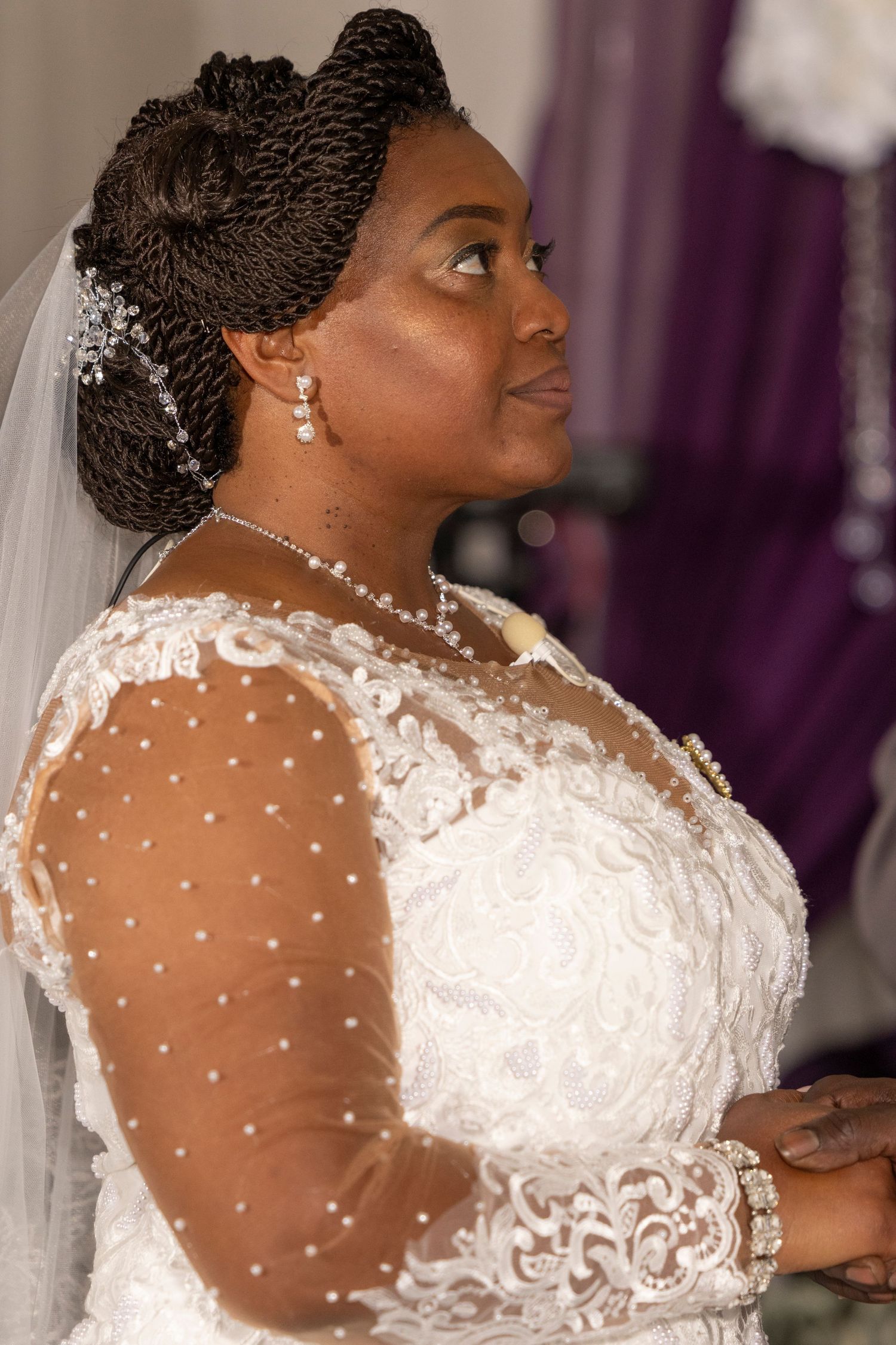 A bride in a white lace gown with pearl details and veil looks up gracefully during her wedding ceremony.