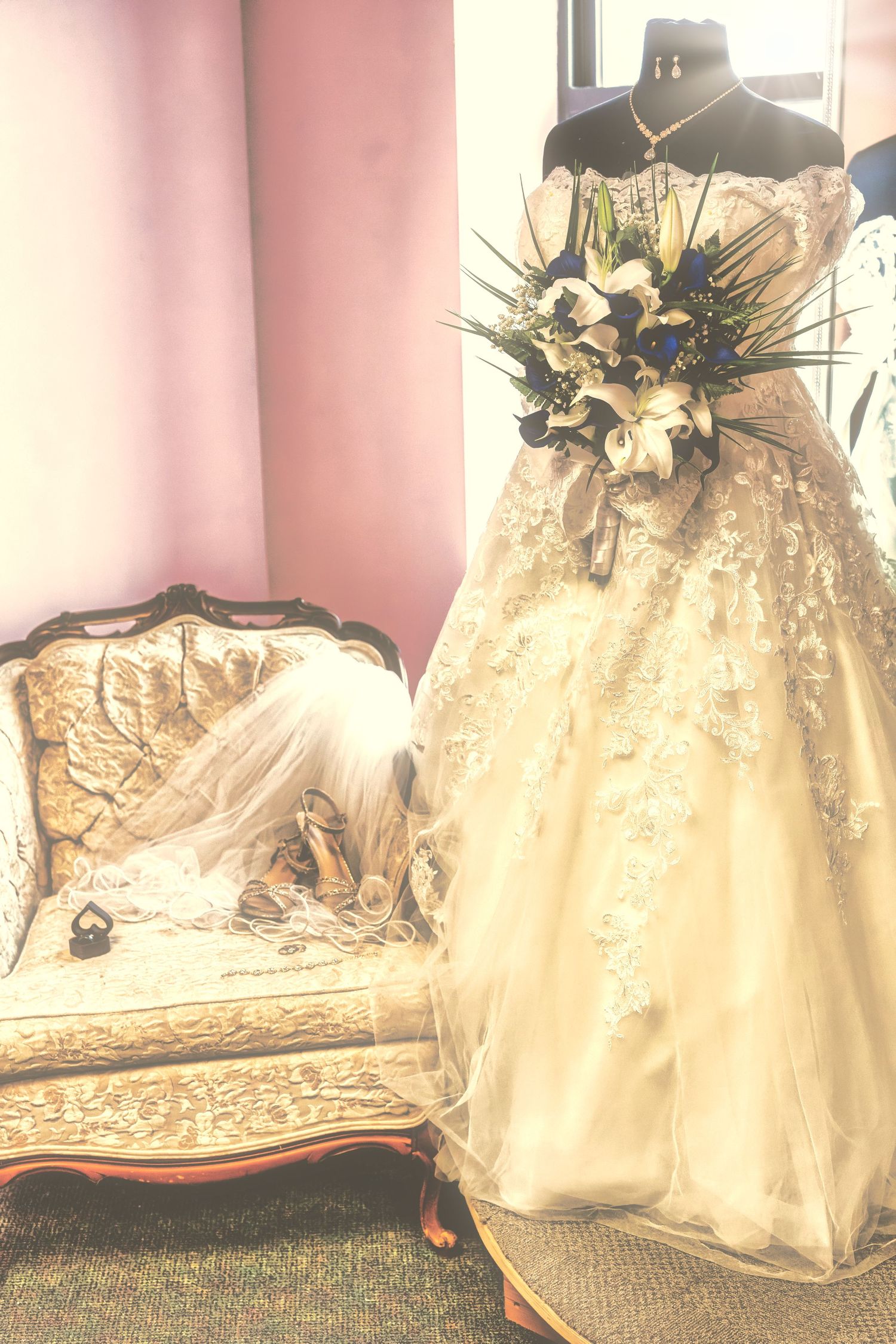 Elegant lace wedding gown on mannequin with floral bouquet beside vintage ornate settee in bridal boutique.