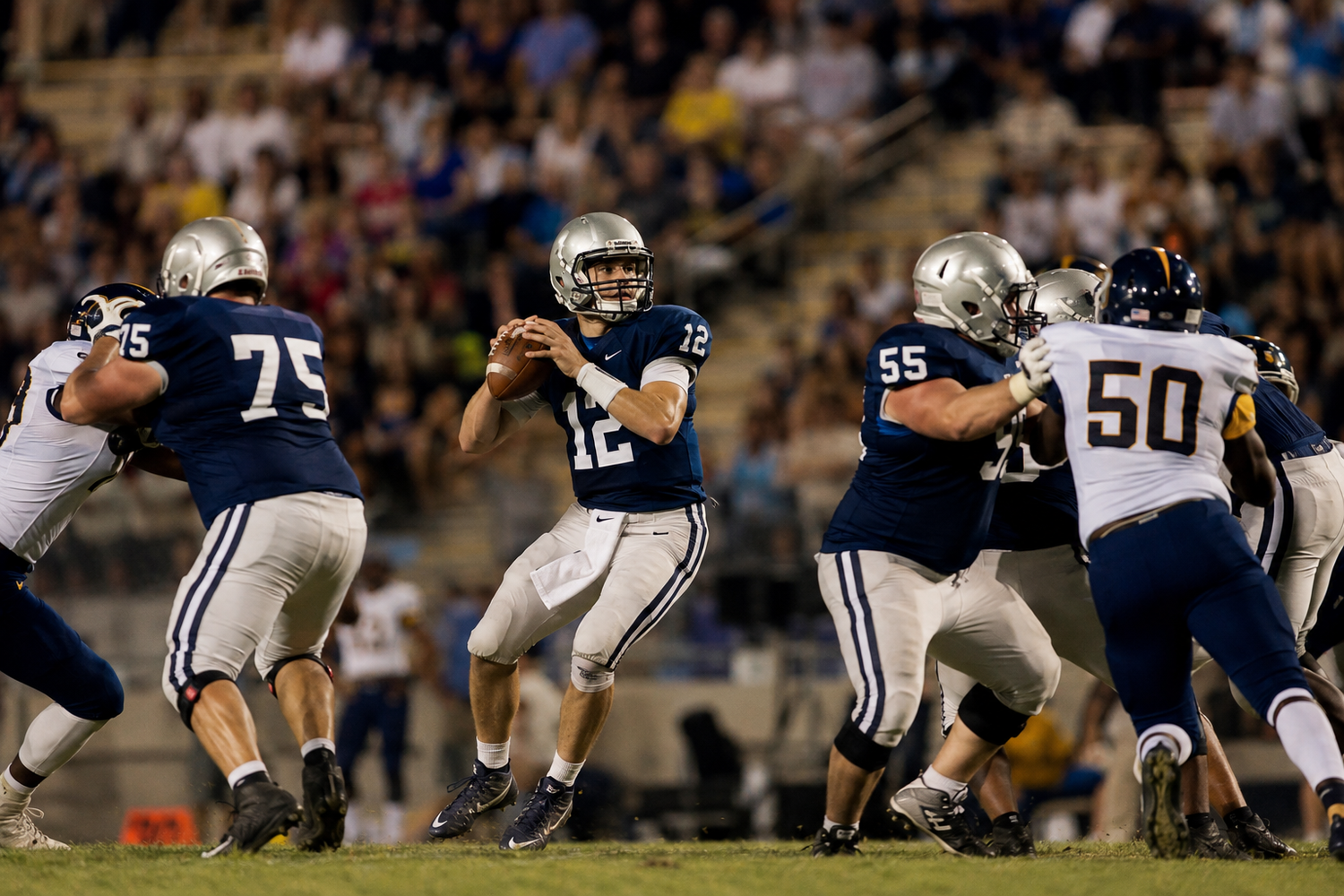 Football quarterback in gray jersey number 12 drops back to pass behind offensive linemen during a night game.