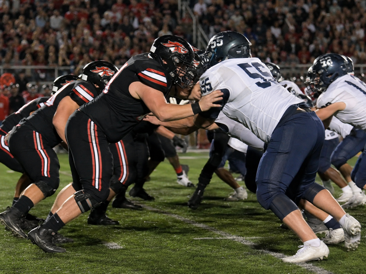 High school football players clash at the line of scrimmage during an intense night game under stadium lights.