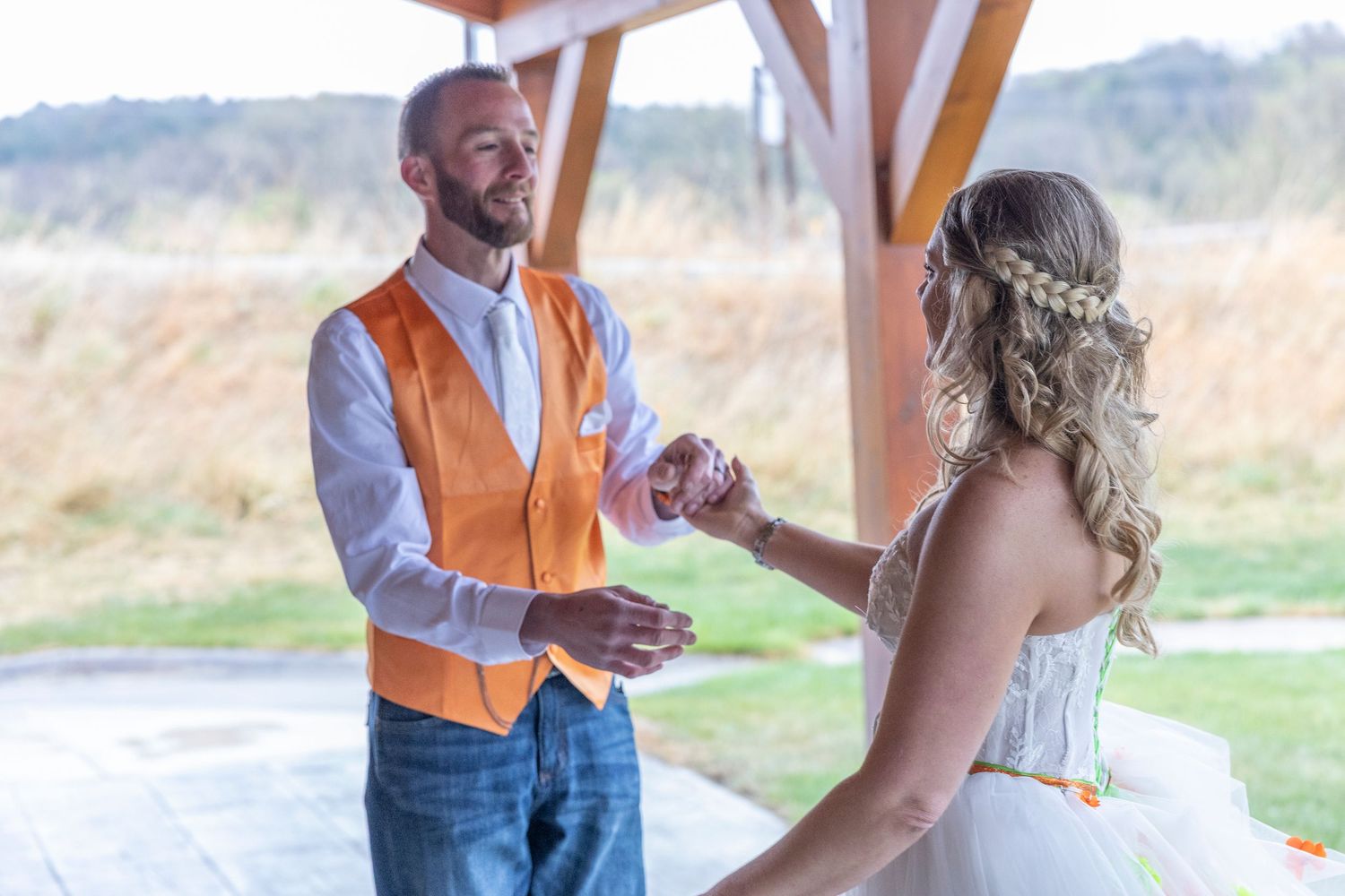 Groom in orange vest and jeans holds bride's hands during outdoor wedding ceremony under wooden pavilion.