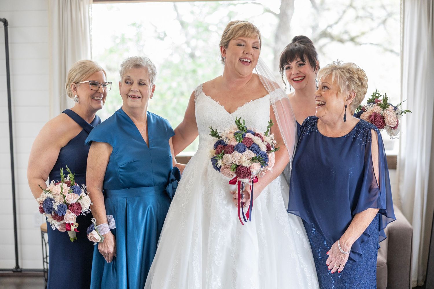 Bride in white gown laughing with four women in blue dresses holding floral bouquets at a wedding celebration.