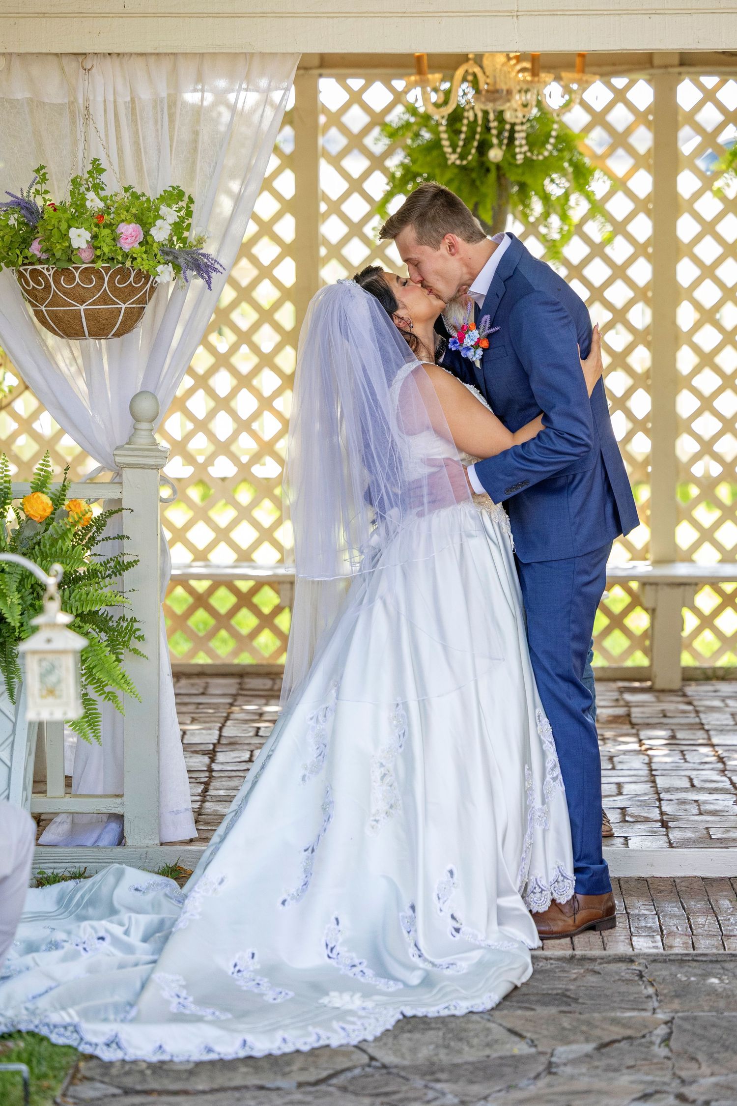 Bride and groom share their first kiss under a decorated gazebo, bride in white gown with veil, groom in navy suit.