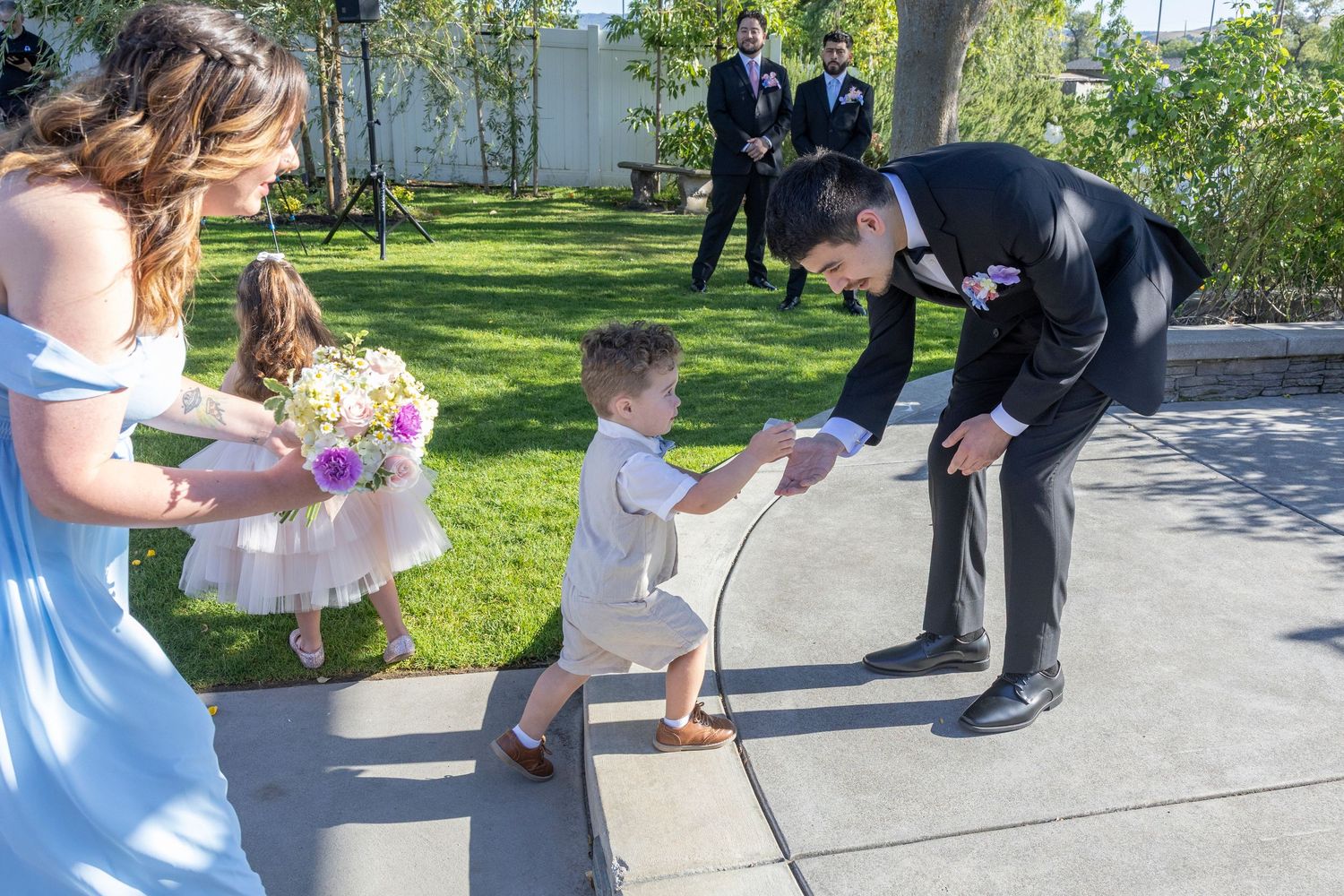 Groom in black suit bends down to greet young ring bearer boy at outdoor wedding ceremony with bridesmaid nearby.