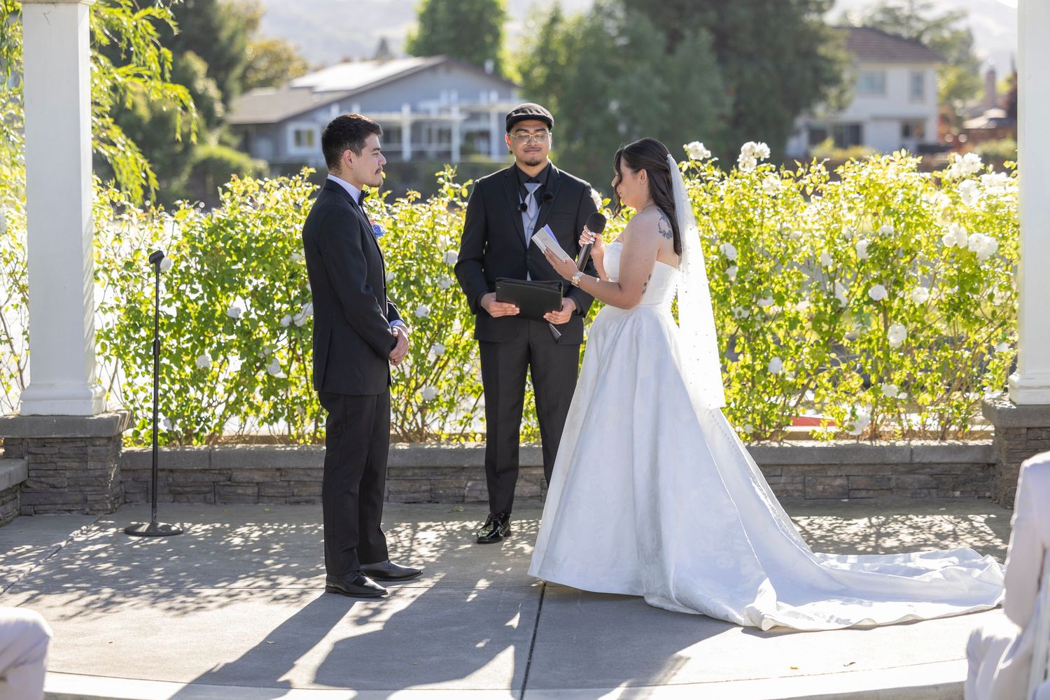Bride and groom exchange vows outdoors with officiant, surrounded by lush greenery at a sunny garden wedding ceremony.