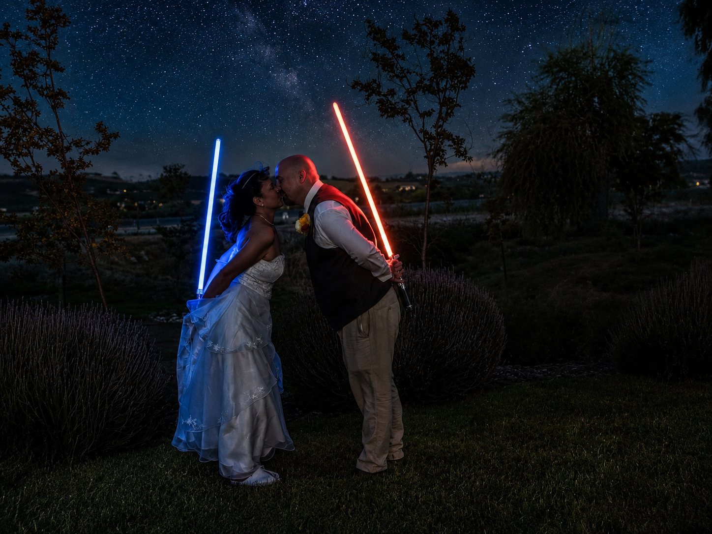 Star Wars themed wedding couple kissing under night sky holding blue and red lightsabers in outdoor ceremony.