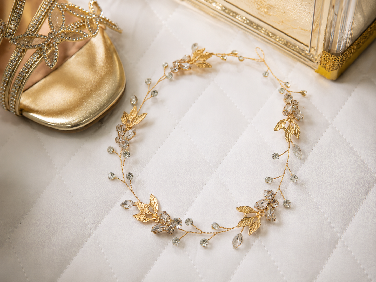 Gold leaf crystal bridal hair vine wreath on white quilted fabric with gold heels and decor in background.