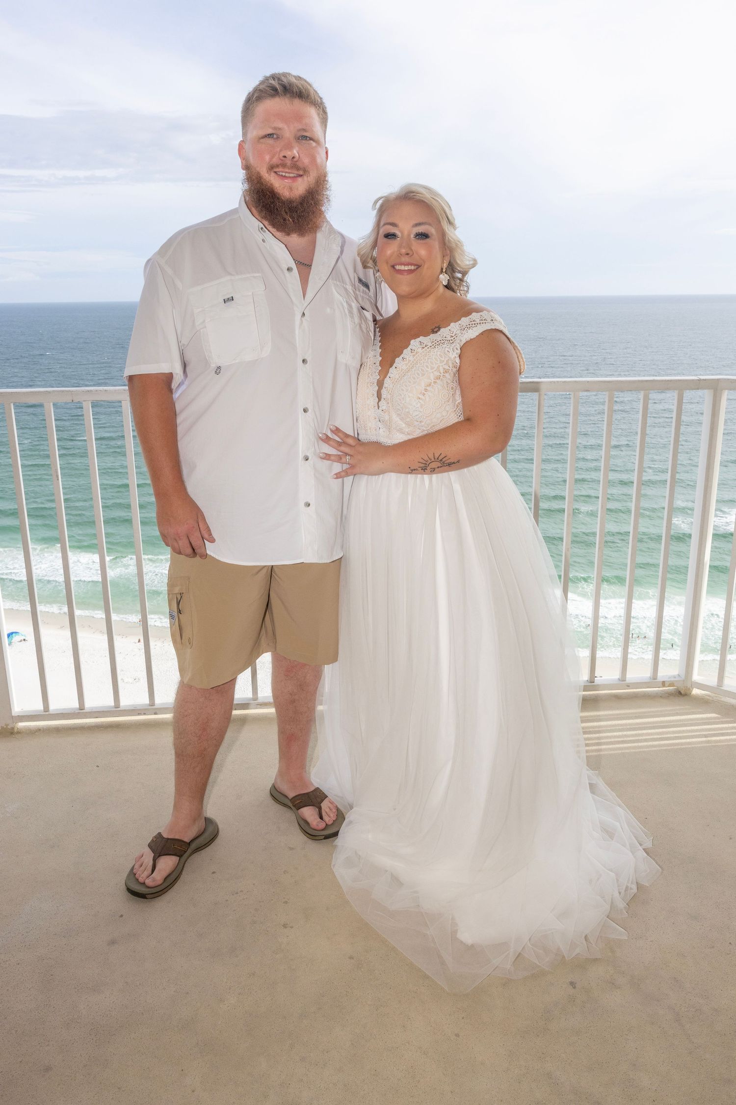 Couple in wedding attire posing on oceanfront balcony, bride in white gown, groom in white shirt and khaki shorts.