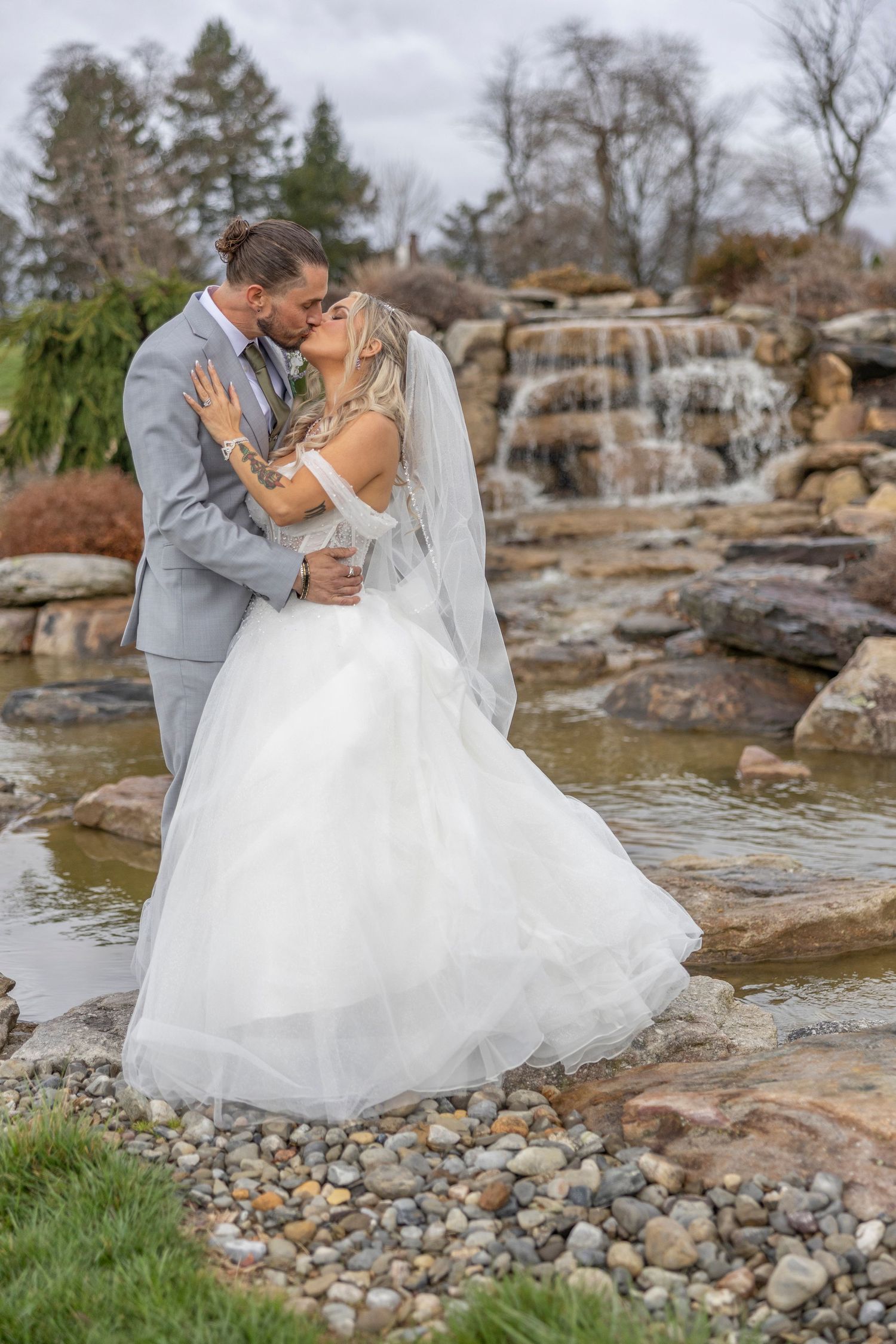A romantic wedding couple embraces by a cascading waterfall on rocky terrain surrounded by evergreen trees.
