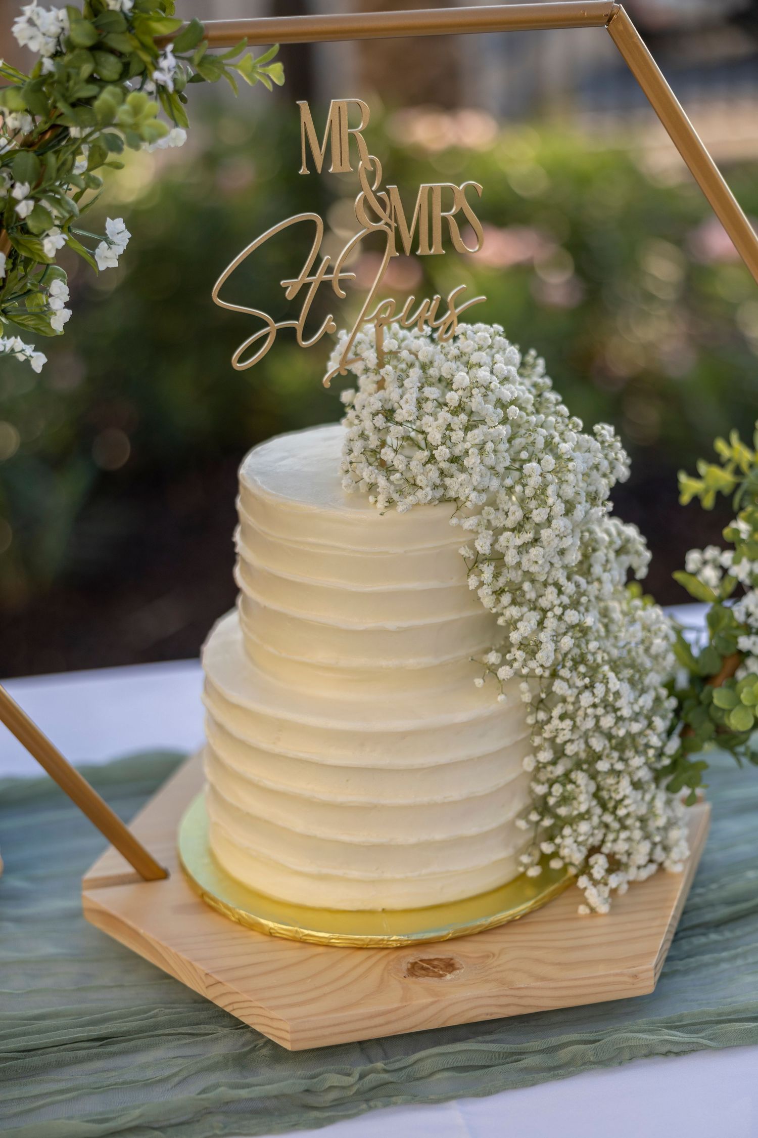 Elegant white wedding cake decorated with baby's breath flowers and gold Mr & Mrs topper on wooden hexagonal stand.