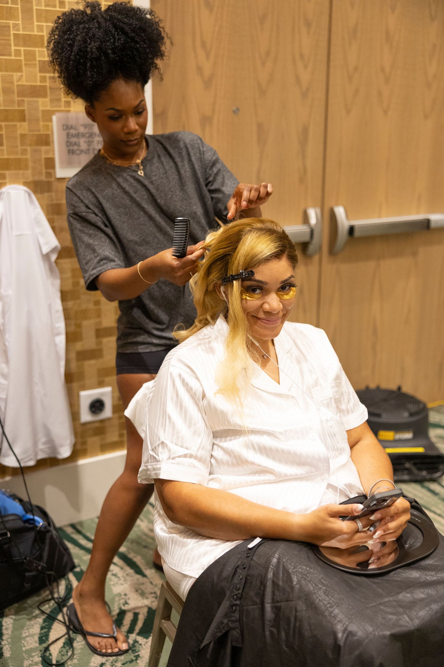 A hairstylist works on styling a client's hair while both check their mobile phones in a salon setting.