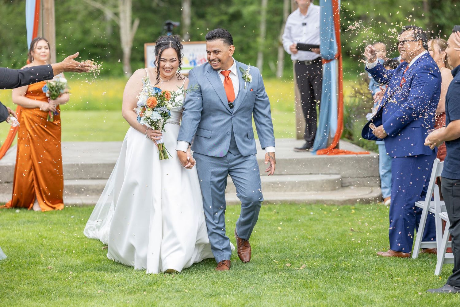 Wedding guests toss rose petals as a newlywed couple walks down an outdoor pathway during a joyful celebration.