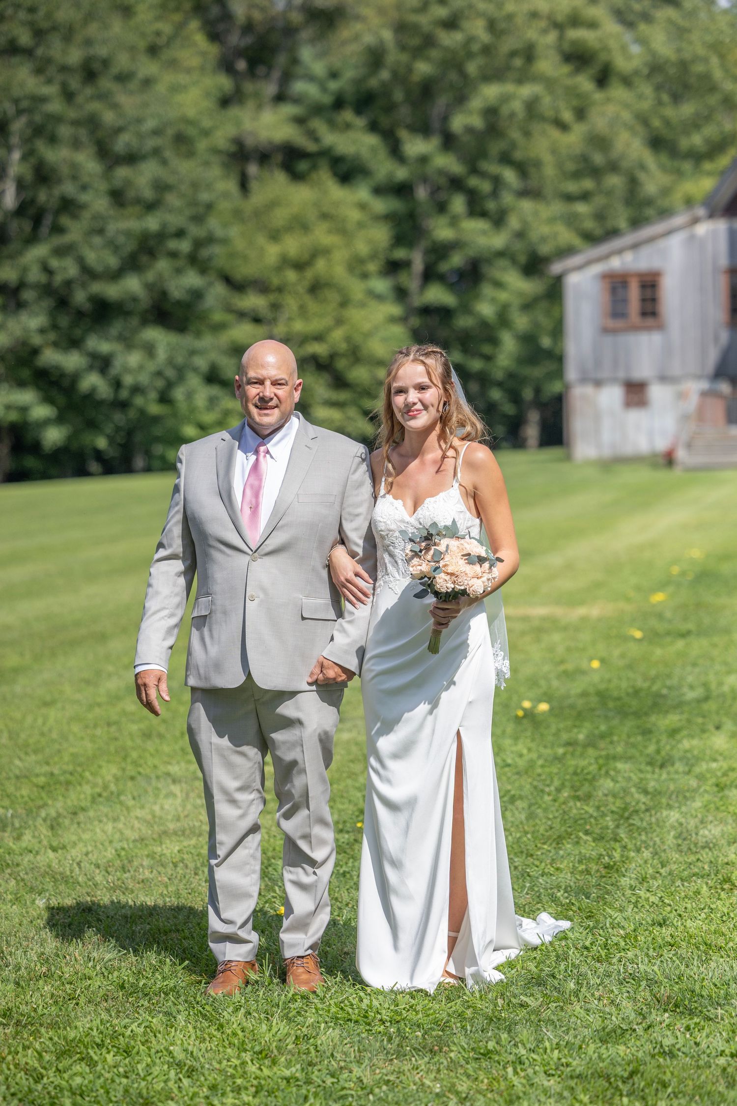 A couple in wedding attire walks together on a green lawn with trees and a rustic barn in the background.