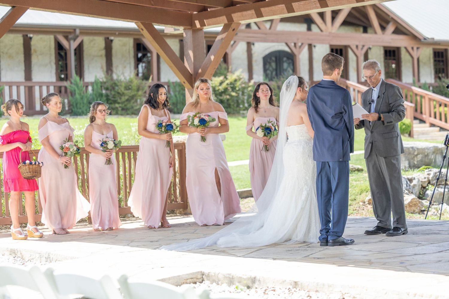 A wedding ceremony takes place under a wooden pavilion with attendants in pink dresses standing in a row.